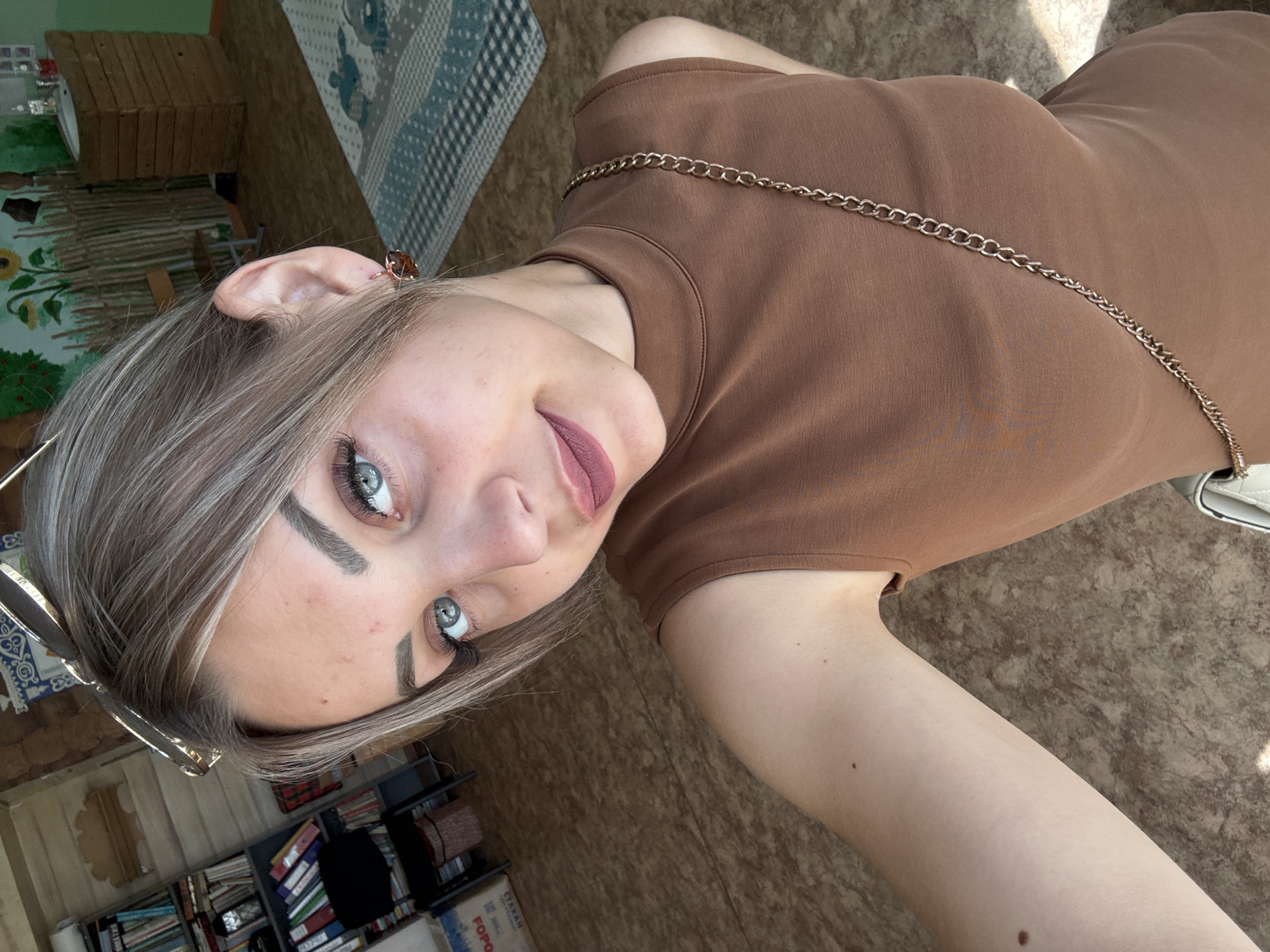 A young Caucasian woman with light brown hair styled in subtle blonde highlights, blue eyes, and fair skin is taking a selfie. She is wearing a sleeveless brown mock-neck top and a gold chain strap is visible across her chest. She has on makeup, including eyeshadow, mascara, and a matte lipstick in a muted berry shade. She is also wearing a single earring with a brown stone in her right ear and sunglasses are perched on top of her head. The background suggests an indoor setting, possibly a room in a school or community center, with a wall mural featuring sunflowers and a wooden fence. To the left, a bookshelf filled with books and binders is visible, along with a chair and a woven basket. A portion of a patterned rug is on the floor. The overall lighting indicates daylight, possibly from a window. The location is identified as Yuzhno-Sakhalinsk, Russia.