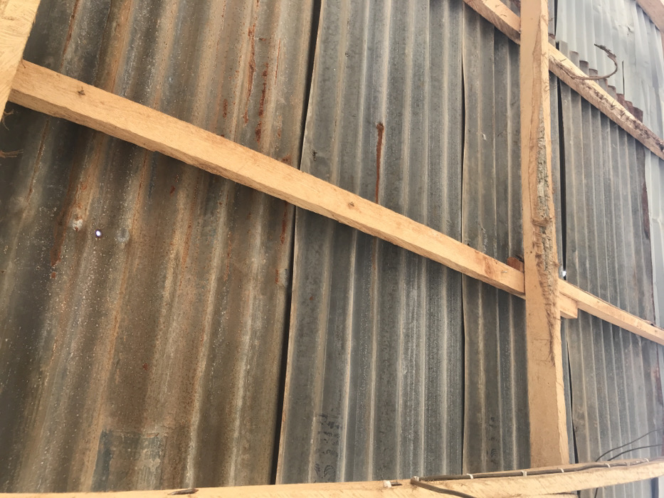 The image displays the underside of a roof structure situated in Oba-Ile, Nigeria. No individuals are present. The primary roofing material consists of corrugated metal sheets, exhibiting a mottled grey and oxidized reddish-brown coloration from extensive rust and weathering. These sheets are supported by an exposed framework of light-brown wooden beams, some displaying rough-cut surfaces and splintering. A distinct circular aperture in an upper corrugated sheet allows a visible point of external light to penetrate. Black insulated electrical wiring is secured vertically along a wooden beam on the left side of the frame. A single bent, rusted metallic object, consistent with a hook or fastener, is partially visible in the lower right quadrant.