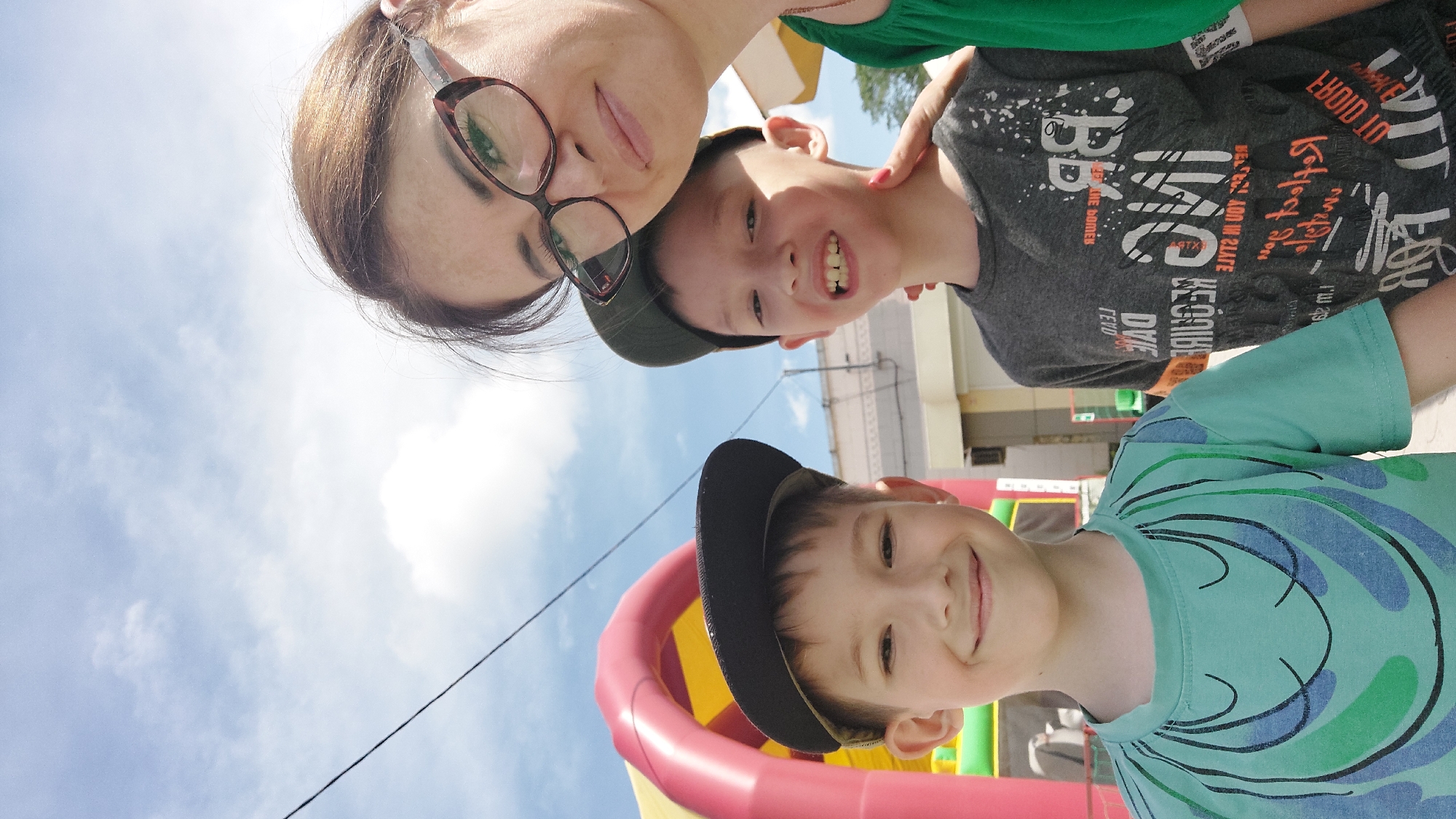 This is a selfie taken outdoors with a bright blue sky and scattered clouds. A woman wearing glasses and a green shirt is on the right. To her left are two young boys, both wearing baseball caps. The boy in the center has a gray t-shirt with white text. The boy on the left is wearing a teal t-shirt with a smiling face design. Behind them, there is a glimpse of a red and yellow inflatable structure, possibly a bouncy castle. The scene appears to be a daytime outdoor gathering or event. This image was captured in the city of Komsomolsk-on-Amur, Russia.