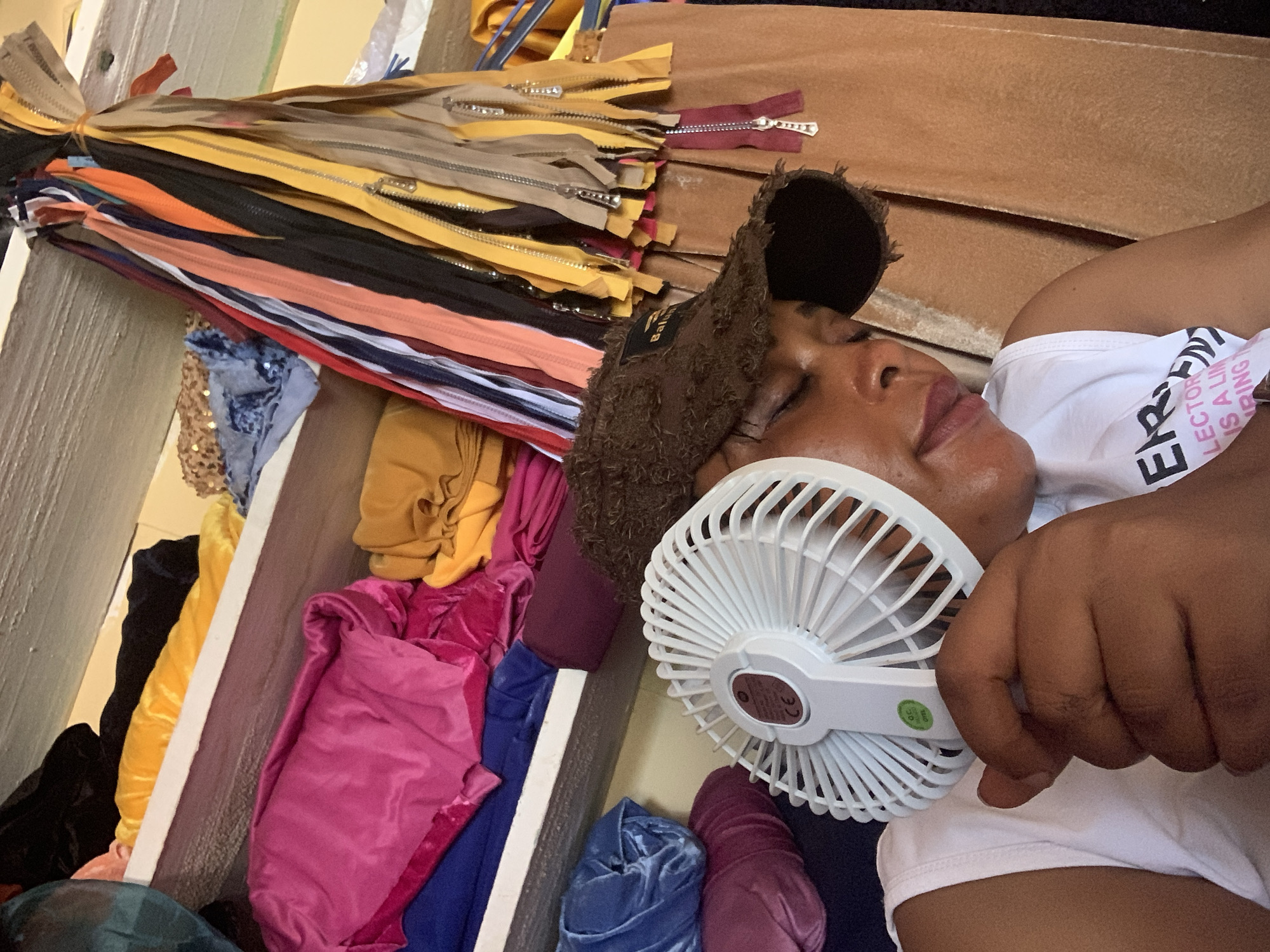 In Yelwa, Nigeria, a woman with dark skin wears a brown baseball cap and a white t-shirt. She is holding a white portable fan close to her face, her eyes are closed. She appears to be resting in a room filled with rolls of fabric and a bundle of colorful zippers hanging from the ceiling. The shelves are stacked with various textiles, including pink, blue, yellow, and maroon velvet materials, as well as a piece of sequined fabric. The environment suggests a workspace or storage area for a tailor or fabric vendor.