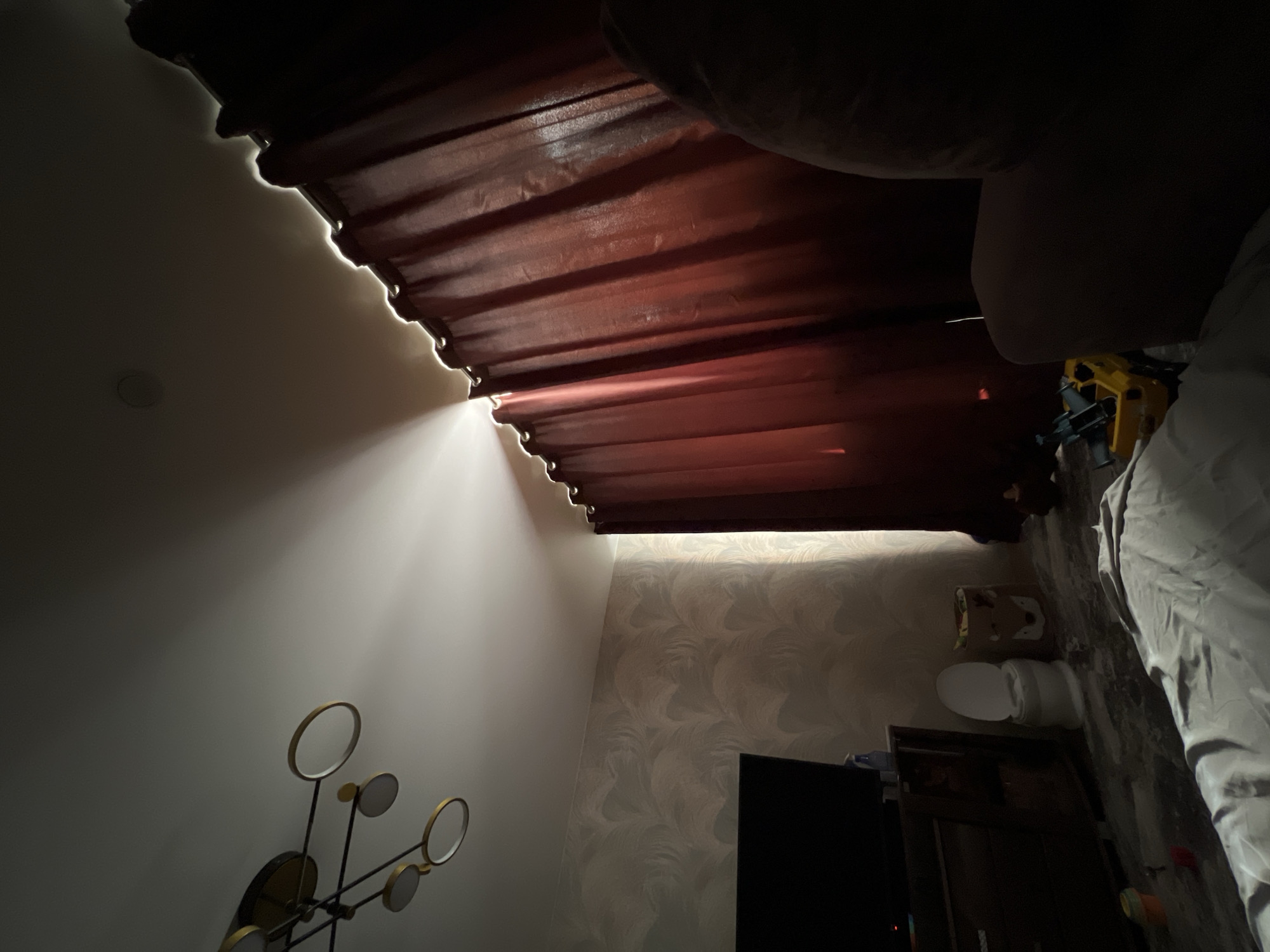 This image presents an indoor scene, likely a room in Noviy, Russia, captured from a low, tilted perspective that gives the impression of the room being rotated. The most prominent feature is a beam of bright natural light streaming into the space from behind a dark red, pleated curtain that hangs across the upper portion of the frame. This sunlight illuminates the left wall, creating a stark contrast with the shadowed areas.
On the lower-left wall, a modern decorative art piece or light fixture with several metallic rings and circular elements is visible. The back wall features a patterned wallpaper with subtle, wavy designs. Below this, a dark, rectangular screen, likely a television, is set on a dark wooden cabinet or piece of furniture. In the bottom right corner, a white toilet is clearly visible against the patterned wallpaper, suggesting either a bathroom or an open-plan studio layout. Next to the toilet, a crumpled white sheet or duvet cover indicates the presence of a bed. Behind this linen, a yellow, industrial-looking object with blue parts is partially seen. The ceiling in the upper-left part of the frame has a round, recessed light fixture.
The strong, directional sunlight implies it is daytime, possibly morning or late afternoon. No individuals are present, and there is no specific activity taking place. No visible text can be identified in the image. The unique angle and the dramatic natural lighting create a contemplative and high-contrast mood within the room.