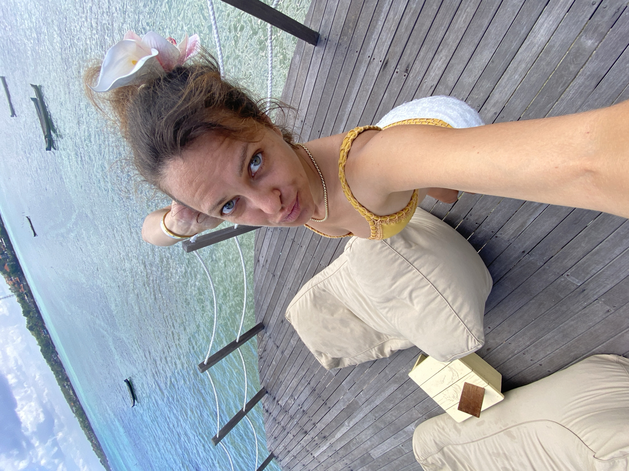 A woman is taking a selfie on a wooden pier in Nusa Penida, Indonesia. She has brown hair with a white flower tucked behind her ear, blue eyes, and is wearing a yellow bikini top and white bottoms. She is leaning on her hand, with her arm bent at the elbow, and her legs are extended. In the background, there is a clear blue ocean with several small boats and a distant coastline with buildings. The pier has a rope railing and two large beige beanbag chairs, one of which she is sitting next to. A small, light-colored wooden table with a dark wooden plaque is situated between the beanbag chairs.