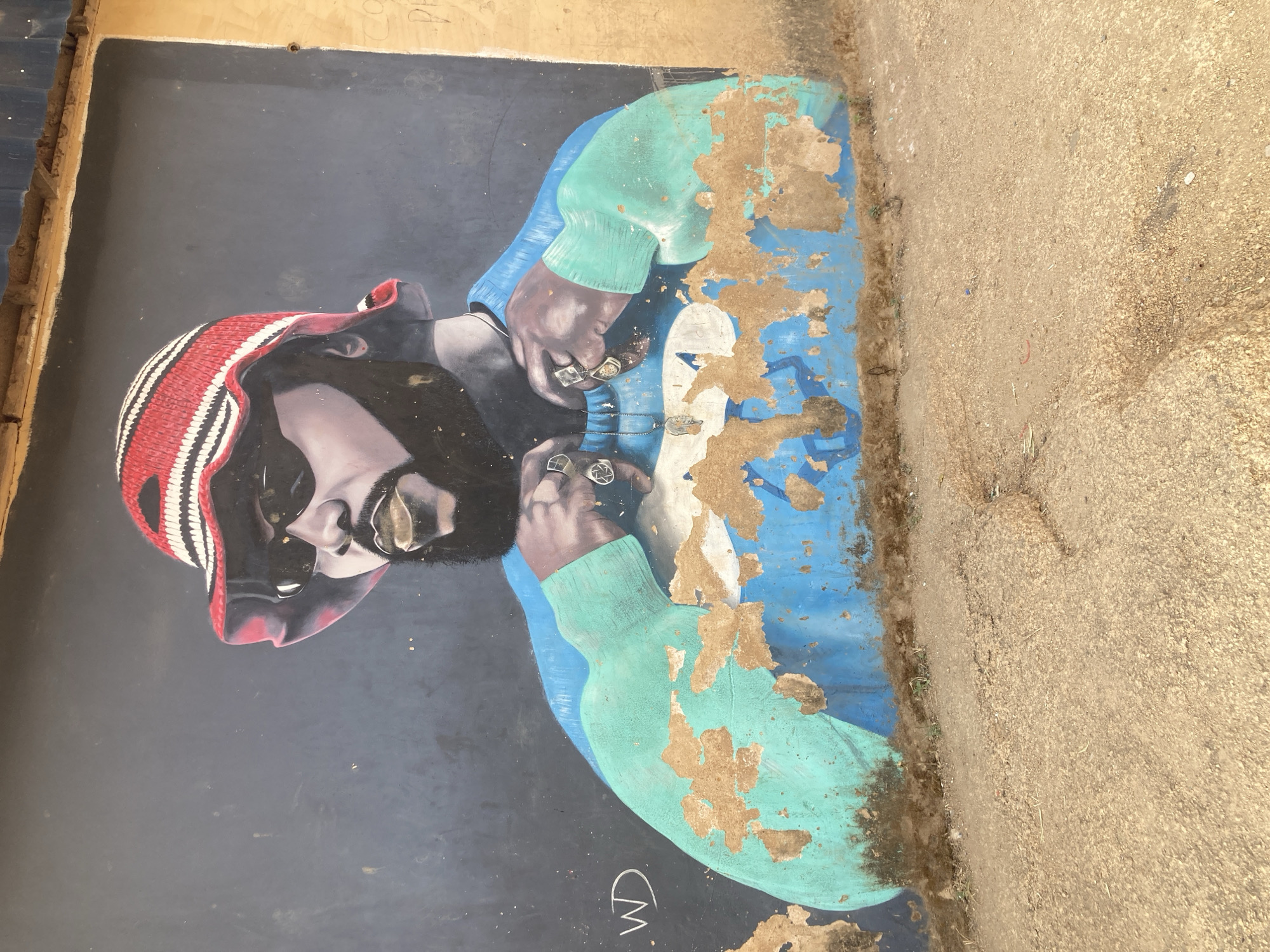 The image shows a mural of a man wearing sunglasses, a red and white striped cap, and a blue and light green jacket. He is depicted fastening a necklace, with rings visible on his fingers. The mural is on a dark surface, likely a wall, and appears to be peeling or damaged in some areas. The location is Jos, Nigeria. A signature "WD" and "CM" is also visible at the bottom left. The mural is positioned next to a rough, concrete-like surface.