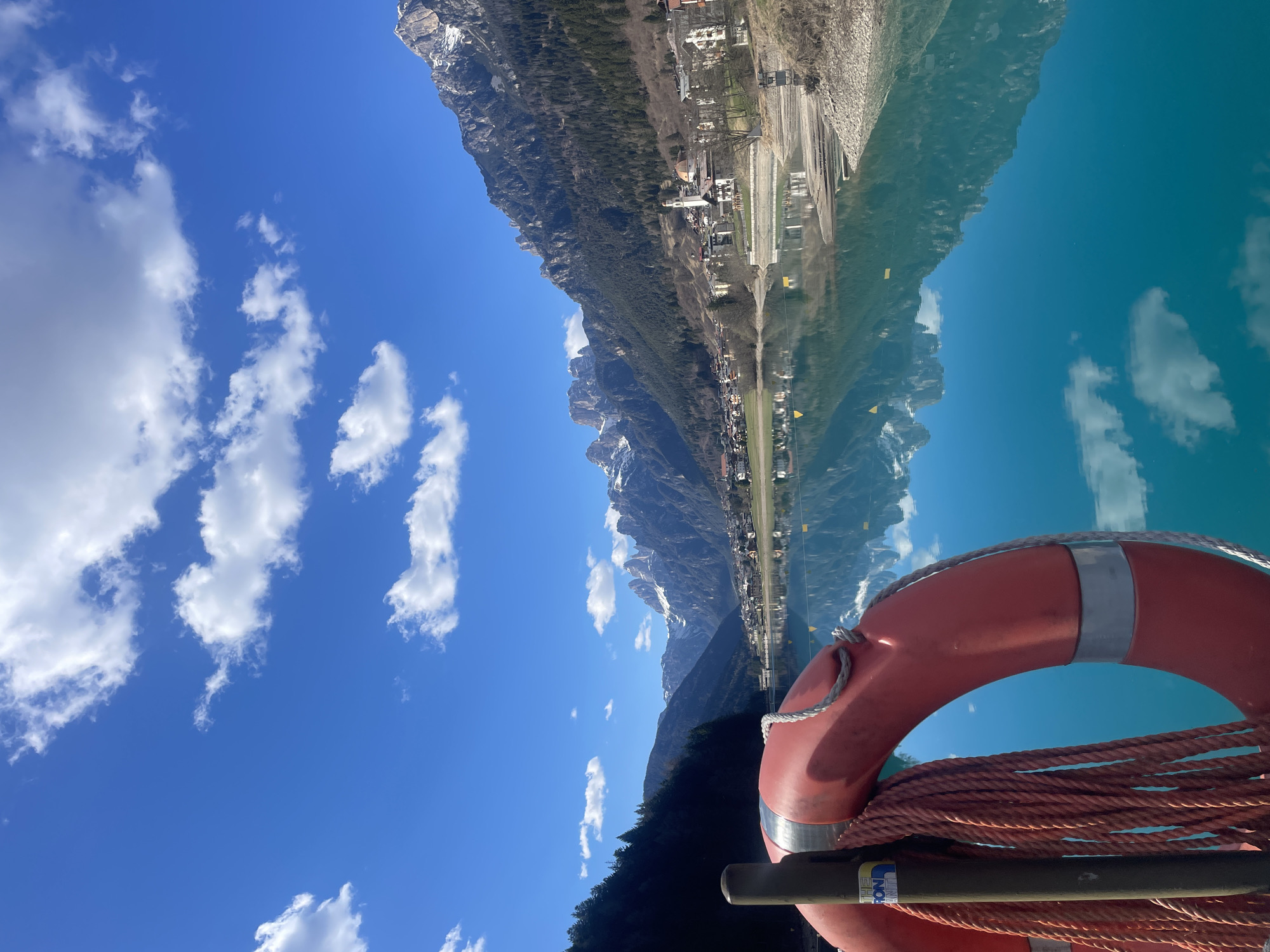 The image shows a view of Auronzo di Cadore, Italy, as seen from a body of water, likely Lake Auronzo. In the foreground is a red life preserver with a rope attached. The lake reflects the surrounding landscape, including the mountains, trees, and buildings of the town. The sky is blue with scattered white clouds.