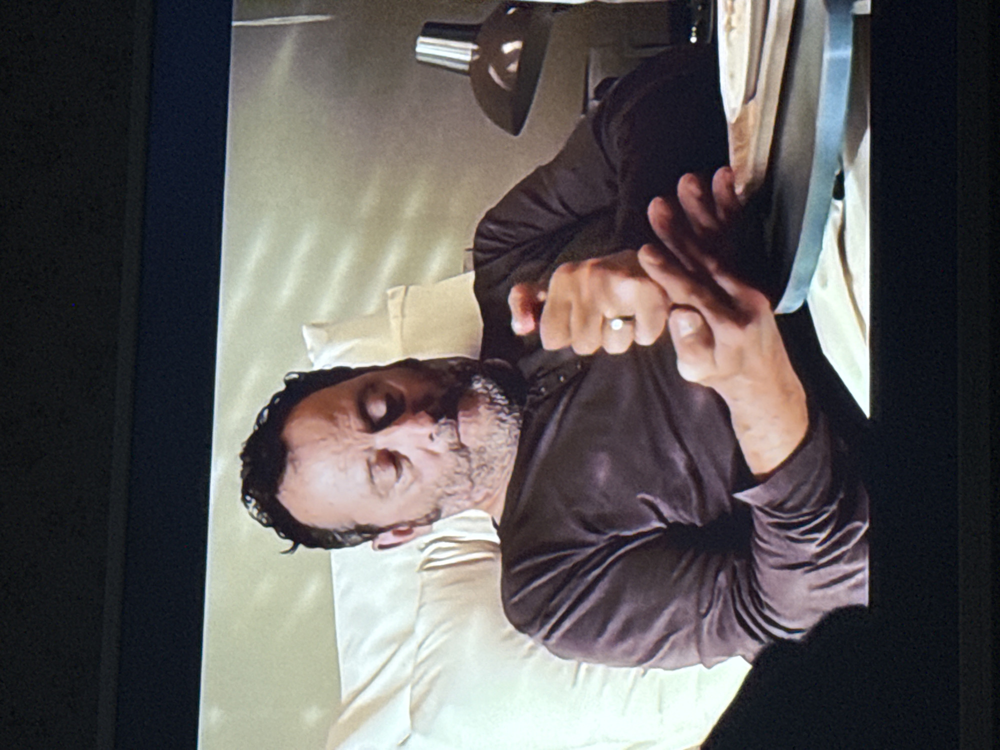 A man with short dark hair and a graying beard lies on a white pillow, his eyes closed. He wears a long-sleeved brown shirt. His hands are clasped together. A metal lamp is positioned above him, and a gray object is next to his hands. The image is taken indoors. There is no evidence or indication of the location of Mytishchi, Russia.