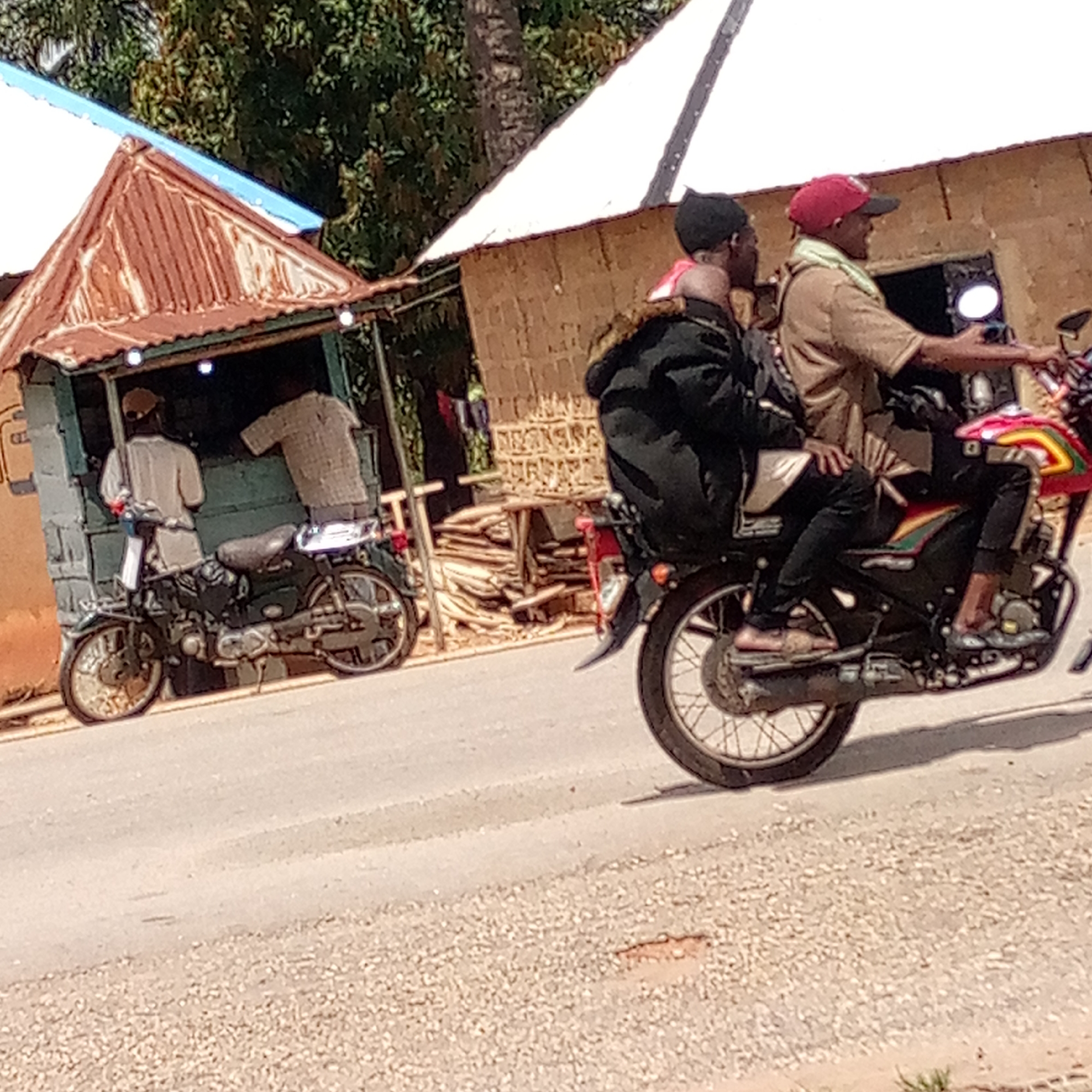 The image shows two men riding a motorcycle on a paved road in Mararaba, Nigeria. The driver is wearing a red cap and light brown shirt, while the passenger is wearing a black hat and black jacket. To the left, there is a small, corrugated metal shop with two men standing near it. A parked motorcycle is positioned in front of the shop. In the background, there is a building with a white roof and a pile of wood.
