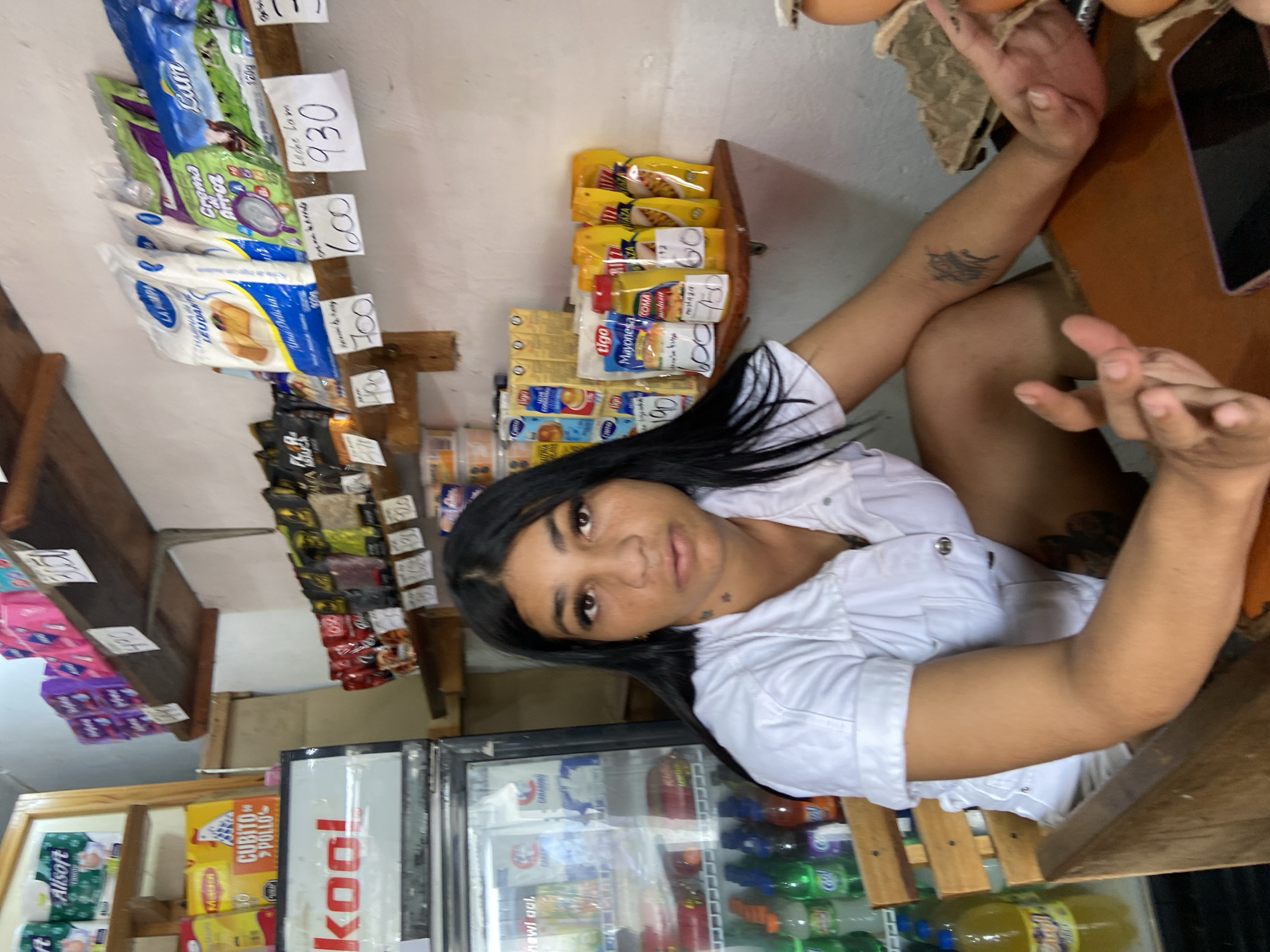 The image captures a medium shot of a woman with dark hair and brown eyes, wearing a white collared shirt, seated at a wooden counter in what appears to be a small convenience store or "bodega." She has visible makeup on and a tattoo on her left forearm. Her hands are resting on the counter. Behind her, shelves are stocked with various food items and household products, including packaged flour, rice, condiments like mayonnaise and mustard, and small containers. To the left, a refrigerated display case contains beverages. The overall environment suggests a local retail setting in San José de Barlovento, Venezuela. The lighting is natural and somewhat dim, consistent with an indoor space during the day.