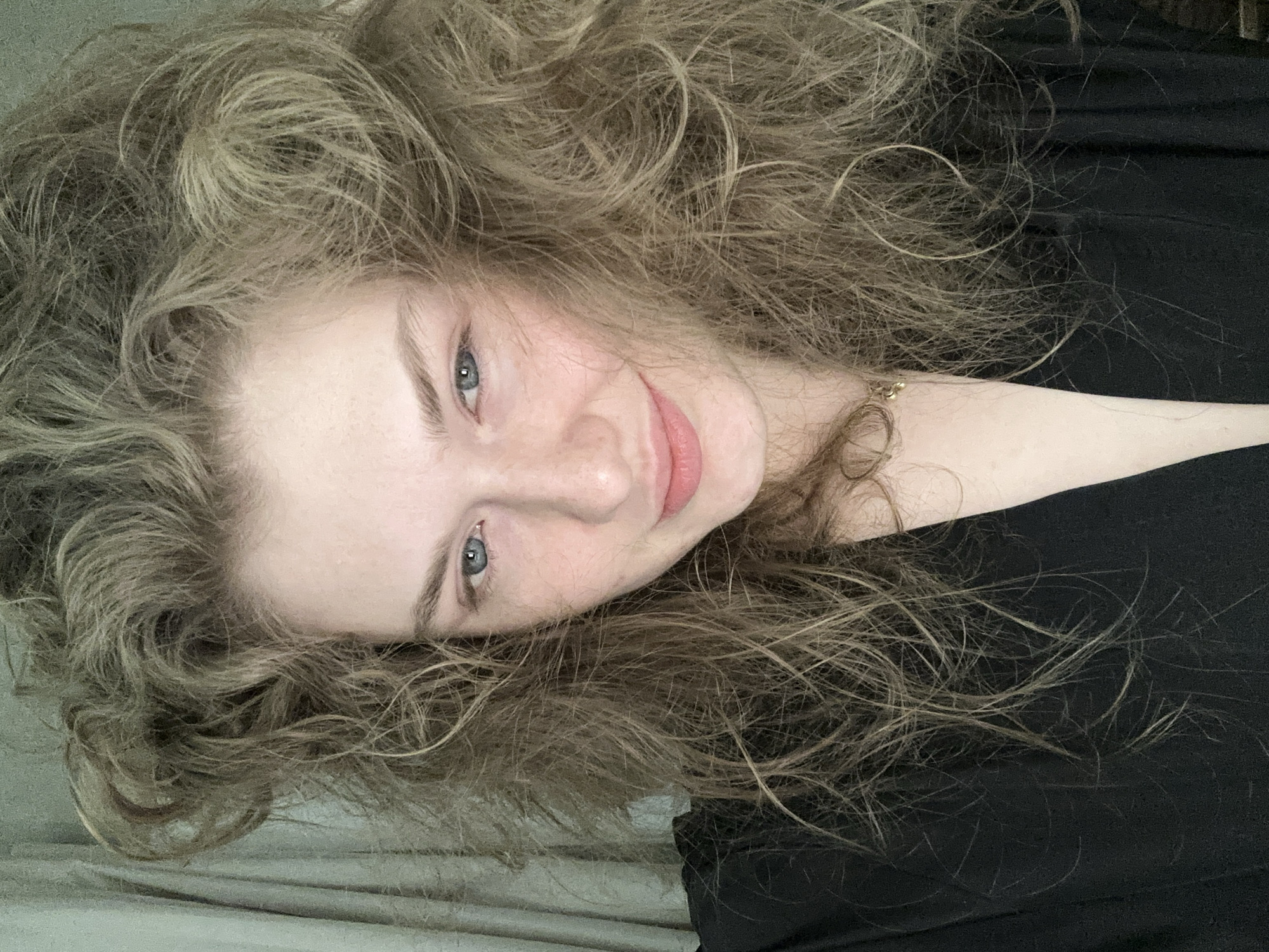 The image depicts a fair-skinned woman with long, wavy, light brown to blonde hair. Her eyes are light blue. She is wearing a black shirt. The background is a light, neutral color, possibly a bed sheet. The woman is positioned on her side, tilting her head towards the camera with a slight smile. There is a gold chain visible at the base of her neck. The image is not taken in the city of Kazan, Russia.
