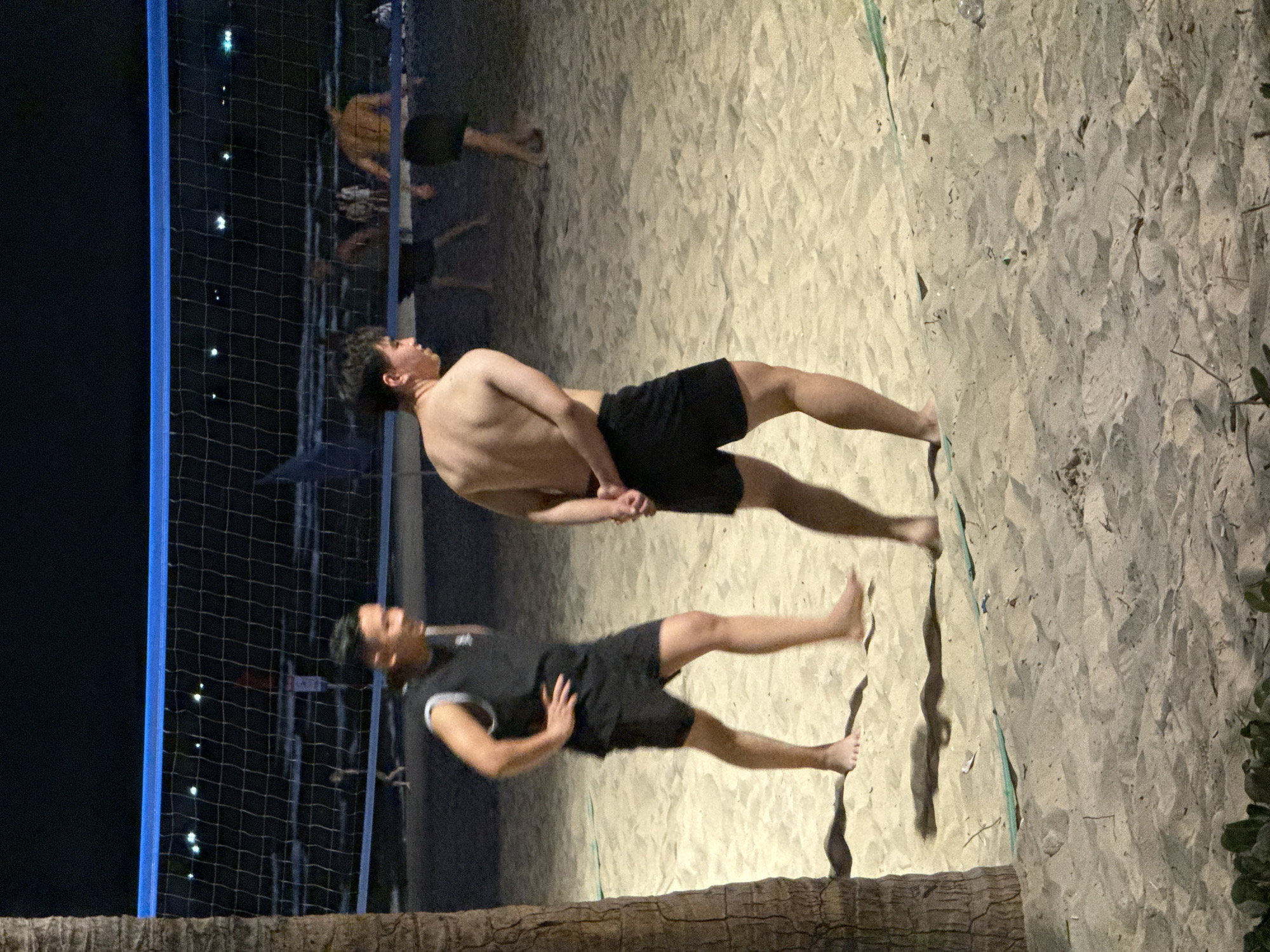 The image shows two shirtless men playing beach volleyball on a sandy beach at night, most likely in Đà Nẵng, Vietnam. One man is wearing black shorts and a sleeveless shirt with a white collar, while the other man is wearing black shorts. There are other figures in the background also appearing to play volleyball. A volleyball net with blue supports is visible on the left. The ground is sandy, and there are some trees or vegetation on the right edge of the frame. The lighting appears to be artificial, illuminating the beach area. A palm tree trunk is visible in the foreground.