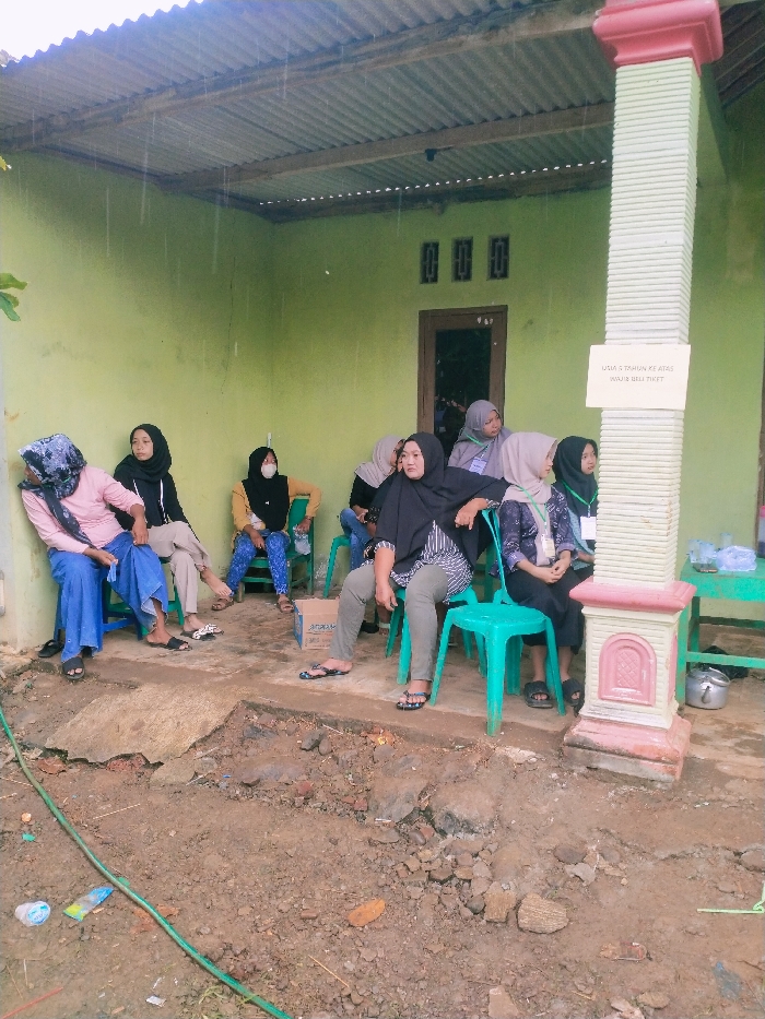 The image shows a group of seven women sitting on chairs under a covered porch in Kecamatan Kedawung, Indonesia (latitude: -7.493, longitude: 111.07). They are all wearing headscarves and traditional clothing. Some women are wearing face masks. One woman is sitting on a blue plastic chair and is wearing a pink shirt and blue pants. Another woman is sitting on a green chair and is wearing a black shirt and brown pants. The woman on the left is holding a handbag. The porch has a green painted wall with a window and three small windows above it. The porch is covered with a corrugated metal roof. The women are sitting in front of a pillar that has a sign that reads "JAMA' S TOHUN KE ATAS BOLEH BILA TIKET." The ground is dirt and there is a green hose running across the ground in the foreground. The scene appears to be informal and casual.
