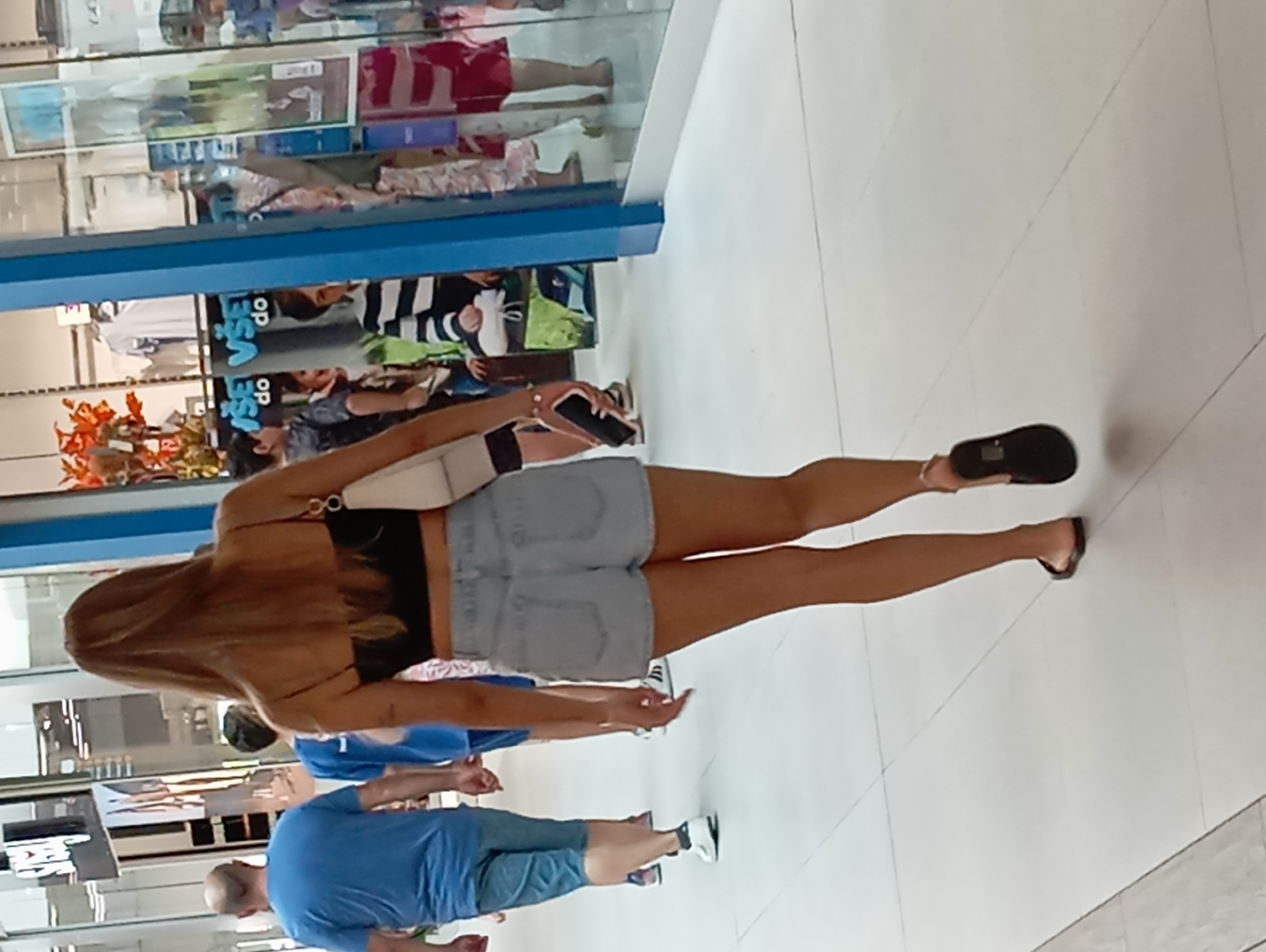This image shows a group of people walking through an indoor shopping mall, possibly in Nitra, Slovakia. In the foreground, a woman with long blonde hair walks away from the camera, wearing a black crop top, denim shorts, and black flip-flops. She is carrying a white purse over her shoulder and holding a smartphone in her right hand. Behind her, a man in a blue t-shirt and blue shorts walks away from the camera, followed by another person in a blue t-shirt. Further in the background, several other individuals, including children, are visible in the mall. The environment is a well-lit, tiled indoor walkway with shop windows visible on the right side of the frame.