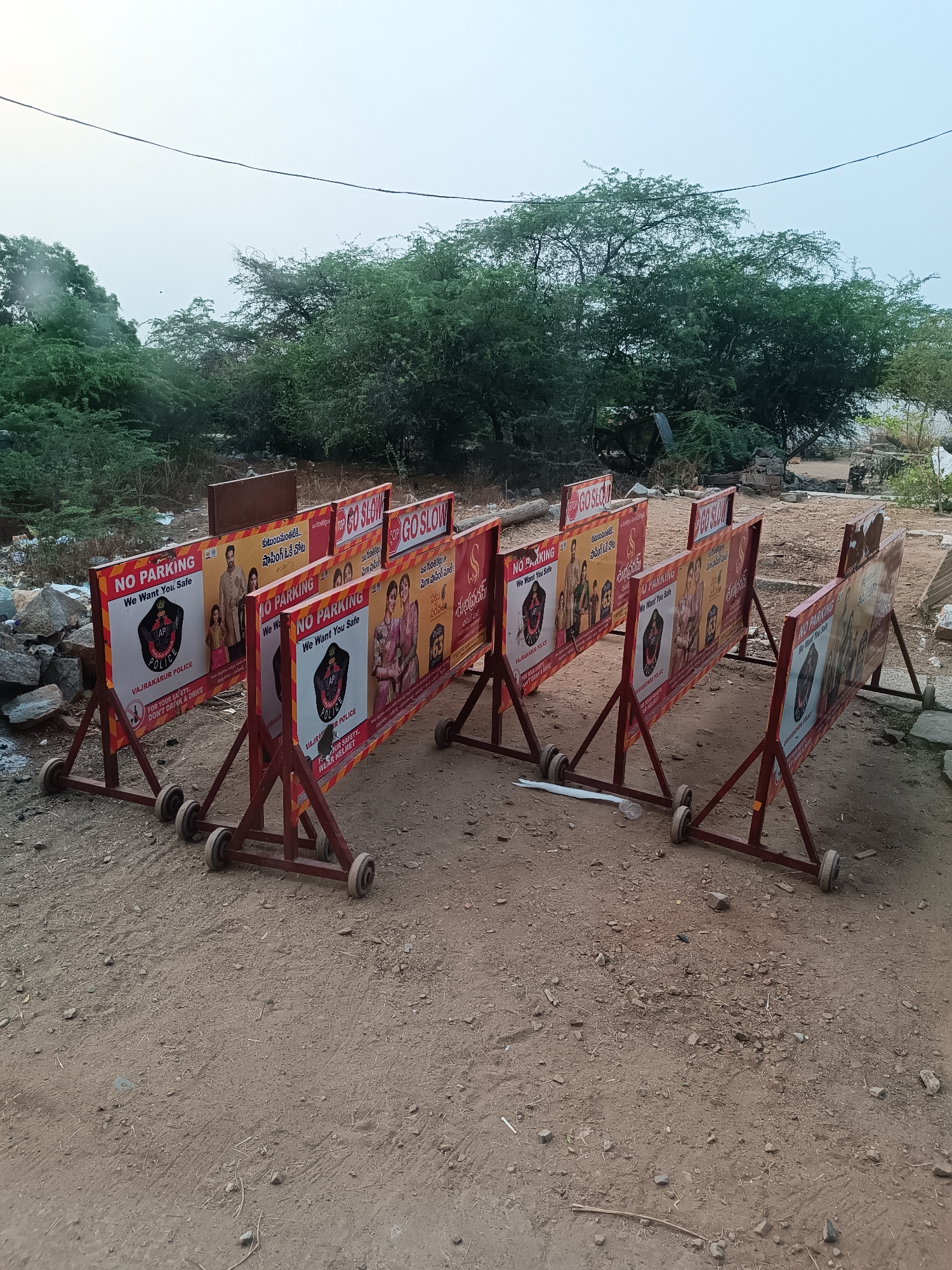 The image depicts a series of six portable, wheeled traffic barriers or signboards arrayed on a dirt path in Chabala, India. The barriers consist of red-painted metal frames supporting rectangular signs. Each sign displays various messages, including "NO PARKING," "GO SLOW," and "We Want You Safe," primarily in English and a local language (likely Telugu). Several signs feature the emblem of "AP POLICE" (Andhra Pradesh Police) or "VAJRAKARUR POLICE," along with images of people demonstrating safe practices like wearing helmets or promoting family safety. The ground beneath the barriers is rough, dry earth interspersed with small stones and sparse dry vegetation. In the background, a dense cluster of green trees and bushes dominates, with a hazy, light-colored sky above. A single power line traverses the upper portion of the frame. No individuals or vehicles are visible in the immediate scene.