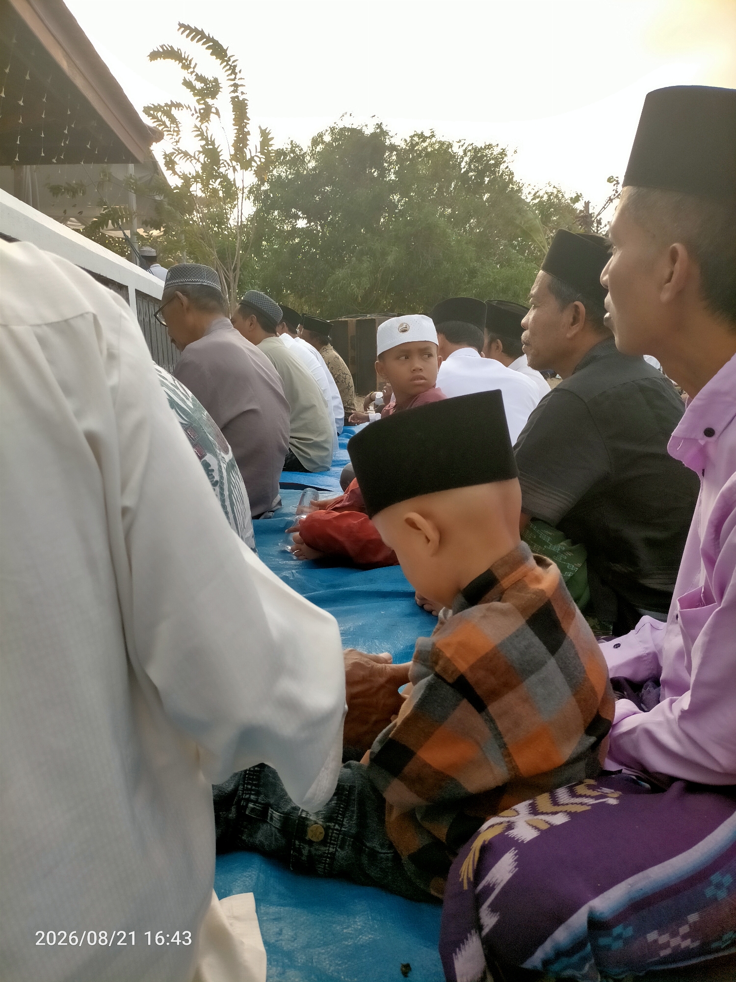 The image depicts a group of people, primarily men and boys, gathered outdoors in what appears to be a community event. They are seated on a blue tarp spread on the ground. Several individuals are wearing white or light-colored traditional Islamic attire, including peci (caps). The background shows trees and a building structure, suggesting an outdoor setting in a populated area, possibly Kranji, Indonesia. The lighting indicates it is daytime, possibly late afternoon due to the warm tones. Some people are looking in different directions, while others appear to be engaged in conversation or prayer. The overall atmosphere seems calm and communal.