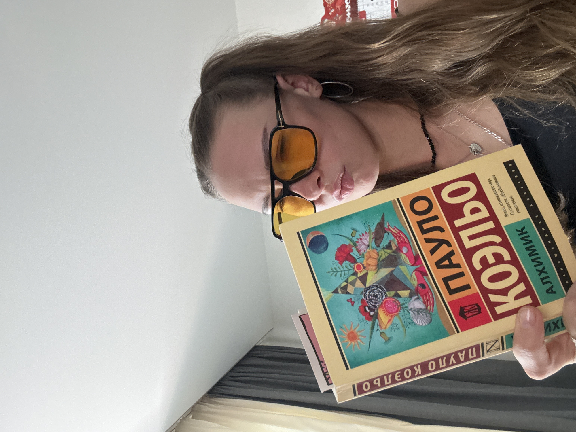 A woman in sunglasses is holding a book in front of her face. The book cover has a colorful illustration with flowers, a sun, a moon, and geometric shapes. Text on the book is in Russian and includes the title "ПАУЛО КОЭЛЬО АЛХИМИК." She is wearing a black top and several necklaces. The background shows a plain white wall and a curtain. The setting appears to be indoors, possibly a room in a residential building.