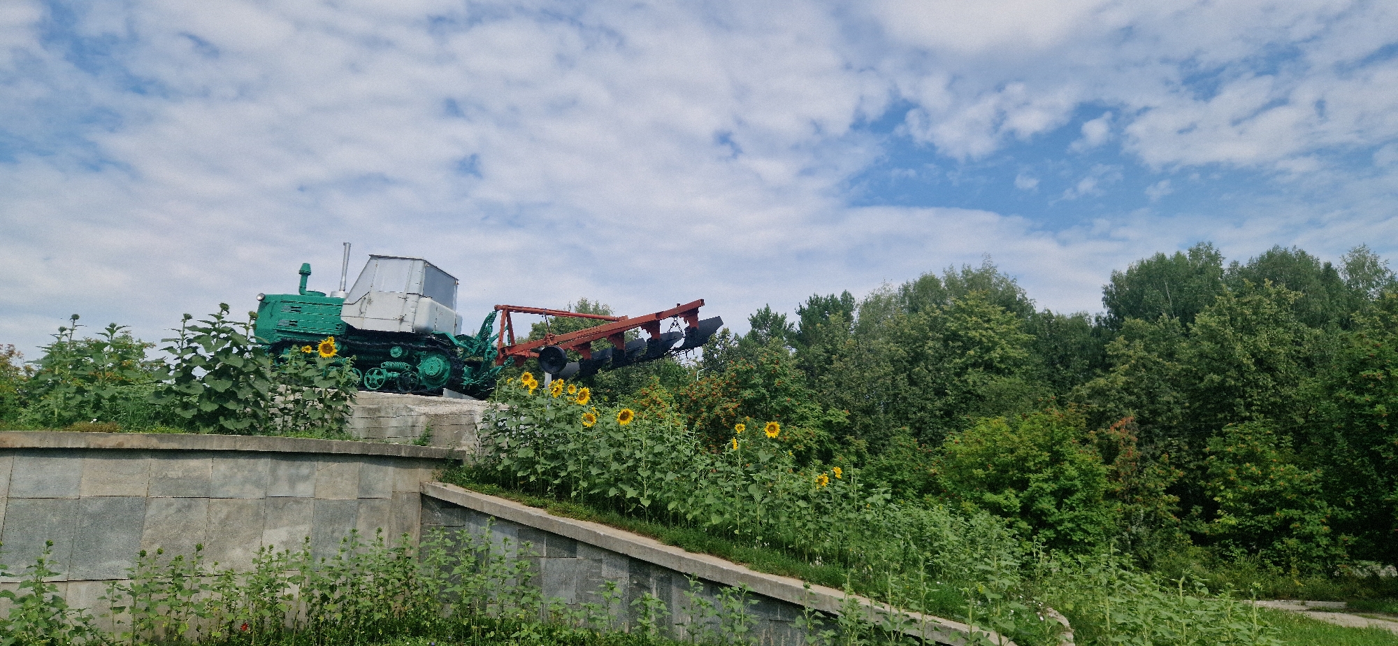 This image depicts a monument to agricultural machinery, specifically a green tractor with a three-bottom plow attached, situated in a park-like setting. The tractor, painted in a distinctive green hue, is positioned on a raised stone base, suggesting it is a permanent display. The plow, a prominent red implement, trails behind the tractor. Surrounding the monument are lush green trees and a field of sunflowers in bloom, adding a vibrant natural element to the scene. The sky above is a clear blue, dotted with scattered white clouds. The overall impression is one of a rural monument commemorating agricultural heritage, likely located in a public space within Krasnoobsk, Russia.