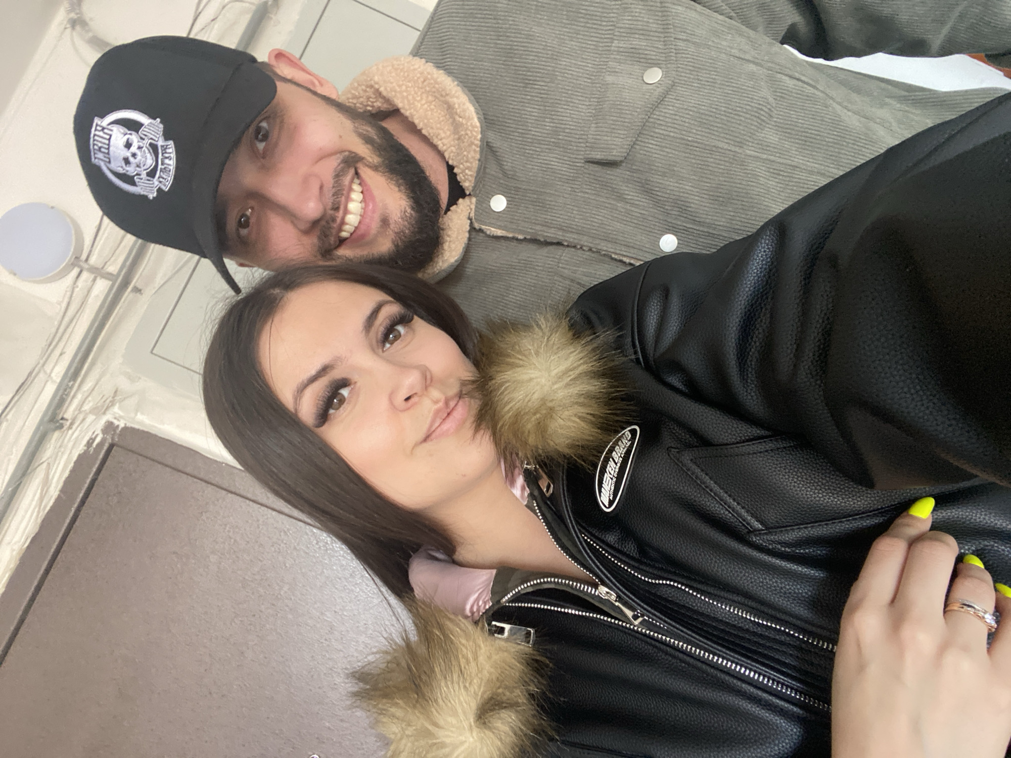 Two people are captured in a selfie. The man is wearing a black baseball cap with a white skull emblem, a gray corduroy jacket with a beige collar, and has a beard. He is smiling and appears behind the woman. The woman is wearing a black leather jacket with fur trim, a light pink top underneath, and has long dark hair. Her fingernails are painted neon yellow. She is wearing a ring. They are positioned in what appears to be a hallway or doorway, with a brown door visible on the left. The light source seems to be artificial and the location is unknown.