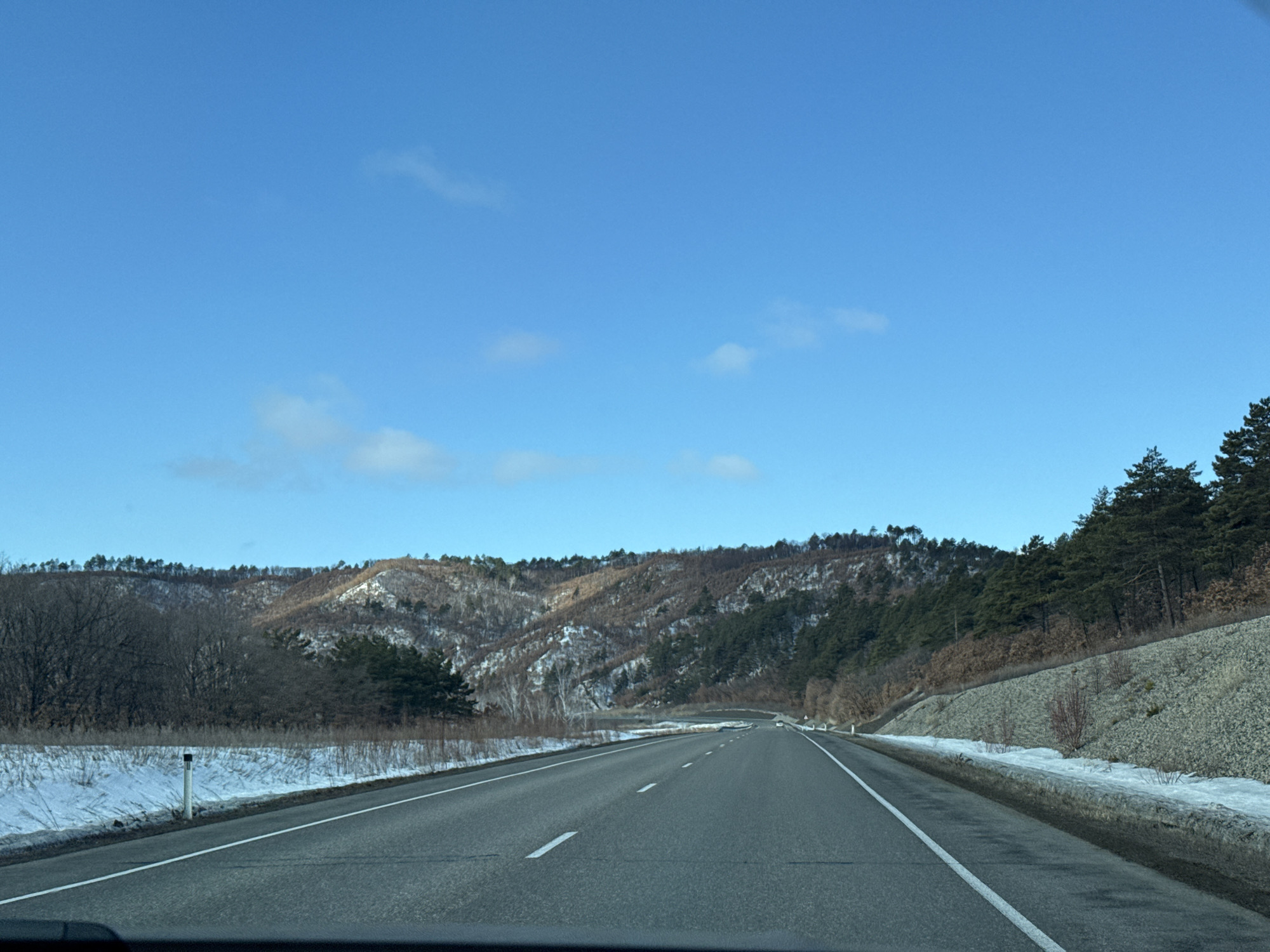 The image displays a two-lane asphalt highway or major road viewed from the interior of a vehicle, looking forward. The road surface is grey with solid white lines on the shoulders and broken white lines for lane division. Patches of snow and dry, brown vegetation cover the ground on both sides of the road. A white marker post with a black cap is visible on the left shoulder. The right side features a gravel embankment leading to a dense stand of evergreen trees. Beyond the immediate roadside, the terrain consists of rolling hills and mountains partially covered in snow. These hillsides exhibit a mix of bare deciduous trees and dark green evergreen trees. The sky is clear and blue with scattered white clouds. Bright daylight illuminates the scene, suggesting a sunny winter or early spring day. No individuals are present. A single vehicle is faintly discernible in the far distance on the highway. The specific city or country (Unknown, Unknown) cannot be determined from the visual evidence; however, the scene depicts a mountainous or hilly rural landscape traversed by a highway.