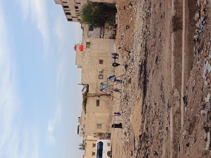 The image depicts an outdoor scene in the Kafr Sousa Municipality, Syria, characterized by a desolate, unpaved urban lot. The foreground is composed of uneven, reddish-brown earth mixed with construction debris, rubble, and scattered refuse, including sections of concrete. In the midground, a rudimentary playground features a blue metal climbing frame or swing set. Several individuals, primarily children, are present near this structure. Specifically, approximately two children are near the blue playground, with one appearing to interact with it. A figure in dark clothing walks towards the right, away from the playground, followed by a small group of children. To the far left, near a black stand-like object, two indistinct figures are visible. Piles of light-colored earth or sand are also scattered across this area. The background features a collection of buildings constructed from light-colored masonry blocks, some appearing complete with windows, while others are partially built or in disrepair. Red water storage tanks are mounted on the rooftops of several completed structures. A white bus or large van is parked on the left side of the background. Sparse green vegetation, including small trees, is visible among the buildings. The sky is light blue with faint, diffuse cloud cover, and a flock of small birds is observed in the upper frame. The lighting suggests daytime conditions.