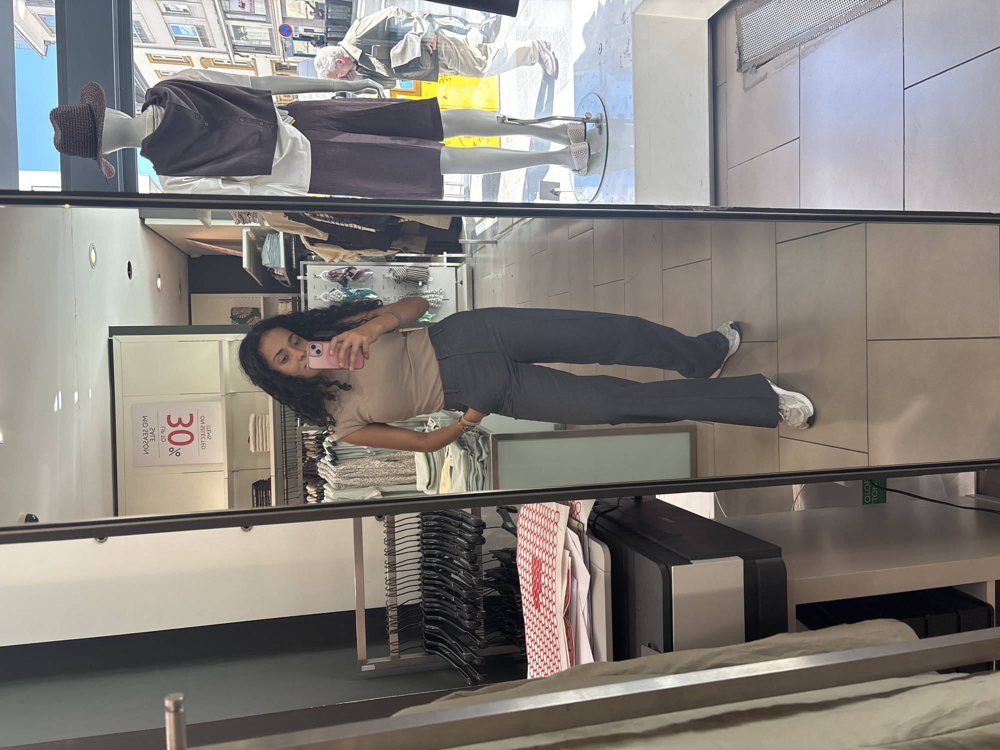 This image is a mirror selfie taken inside a clothing store, capturing a reflection of the person taking the photo along with elements of the store and the outside environment.
The main subject is a young woman with long, dark, curly hair, seen in reflection, holding a pink smartphone to take the picture. She is wearing a light beige t-shirt, dark gray trousers, and white sneakers. Her posture suggests a casual selfie.
The scene includes the interior of the store and a reflection of the street outside. Inside, there are clothing racks with black hangers, shelves displaying neatly folded garments (light blue, white, patterned), and a printer-like device. The store's ceiling is white with recessed lighting, and the floor is tiled in a light, geometric pattern.
In the reflection, two mannequins are visible. One is dressed in a white top, a dark brown cardigan, dark brown bottoms, and a woven brown hat. Only the legs of the second mannequin, wearing light-colored sandals, are seen. Beyond the mannequins and through the store's front window, a sunny, urban street scene is reflected, showing buildings and passersby, including an older man with white hair.
A prominent mirrored sign in the background displays "30%" along with other reversed text that reads "UP TO" and "MID SEASON SALE," indicating a promotional event. The bright light from outside suggests it is daytime with sunny weather. The location is a retail clothing store, consistent with the context of Luxembourg, Luxembourg.