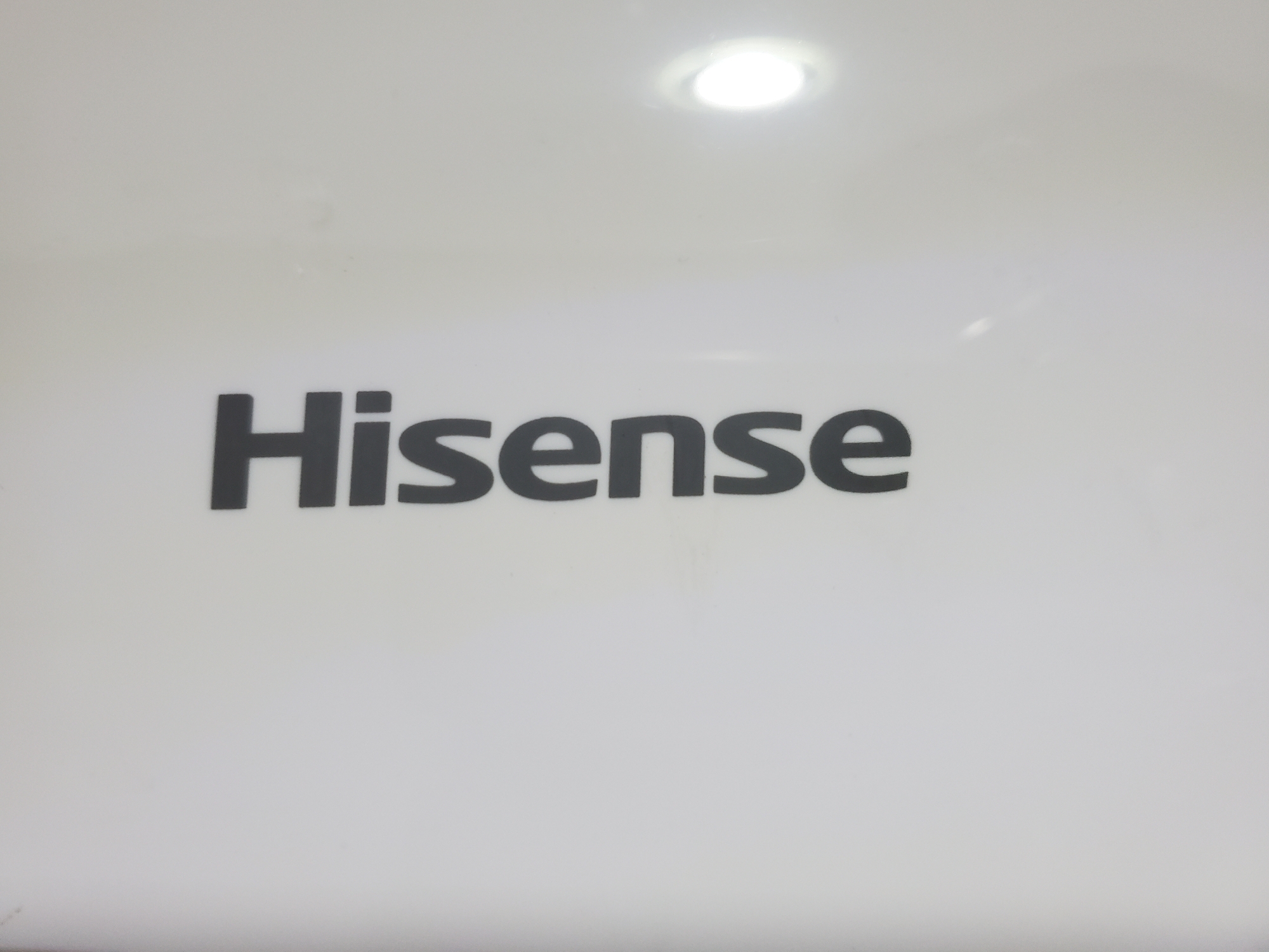 The image is a close-up, eye-level shot of the word "Hisense" in black, bold, sans-serif font, centered on a plain white surface. The surface appears to be a product casing, possibly of an appliance. There is a bright, circular light source reflected in the upper portion of the white surface, casting a slight glare. The overall lighting is even, with no shadows. The background is entirely white and featureless, except for the light reflection. The image does not provide any information about its location in the city of Unknown, Nigeria, or details about people, actions, or interactions.