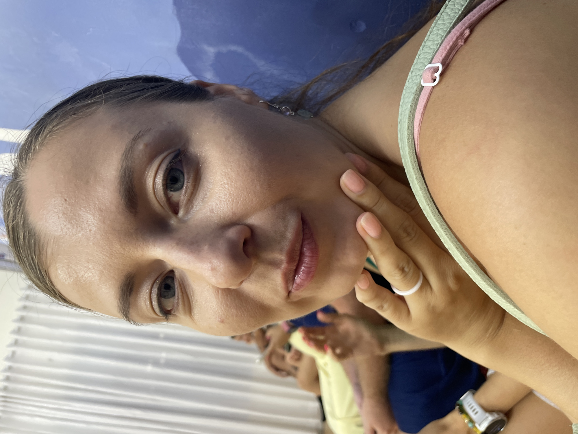 A close-up, portrait-style photograph, originally rotated 90 degrees clockwise, features a young woman looking directly at the camera. She occupies the foreground, positioned slightly to the left, with her head tilted. She has fair skin, light-colored eyes (appears to be blue or grey), and light brown hair that seems to be pulled back. Her lips are a soft pink.
She is wearing a top with visible light green and pink straps. Accessories include a delicate silver earring with a light pink stone and green bead on her left ear, and a white silicone ring on her right ring finger. Her fingernails are painted a light pink. A light-colored smartwatch and a multi-colored beaded bracelet are visible on her right wrist.
Her right hand is gently resting on her jawline/cheek. In the blurred background, several other individuals are partially visible, one wearing a yellow top and another a blue top, suggesting a social gathering or event. The setting appears to be indoors, with a solid blue wall or backdrop behind the woman and white horizontal blinds or louvers visible on the left side of the frame. The lighting is bright and even, consistent with an indoor environment.
The woman exhibits a calm, neutral to slightly pleasant expression with a hint of a soft smile, maintaining direct eye contact. There is no visible text in the image, and no specific time of day or outdoor weather can be determined from the indoor setting.