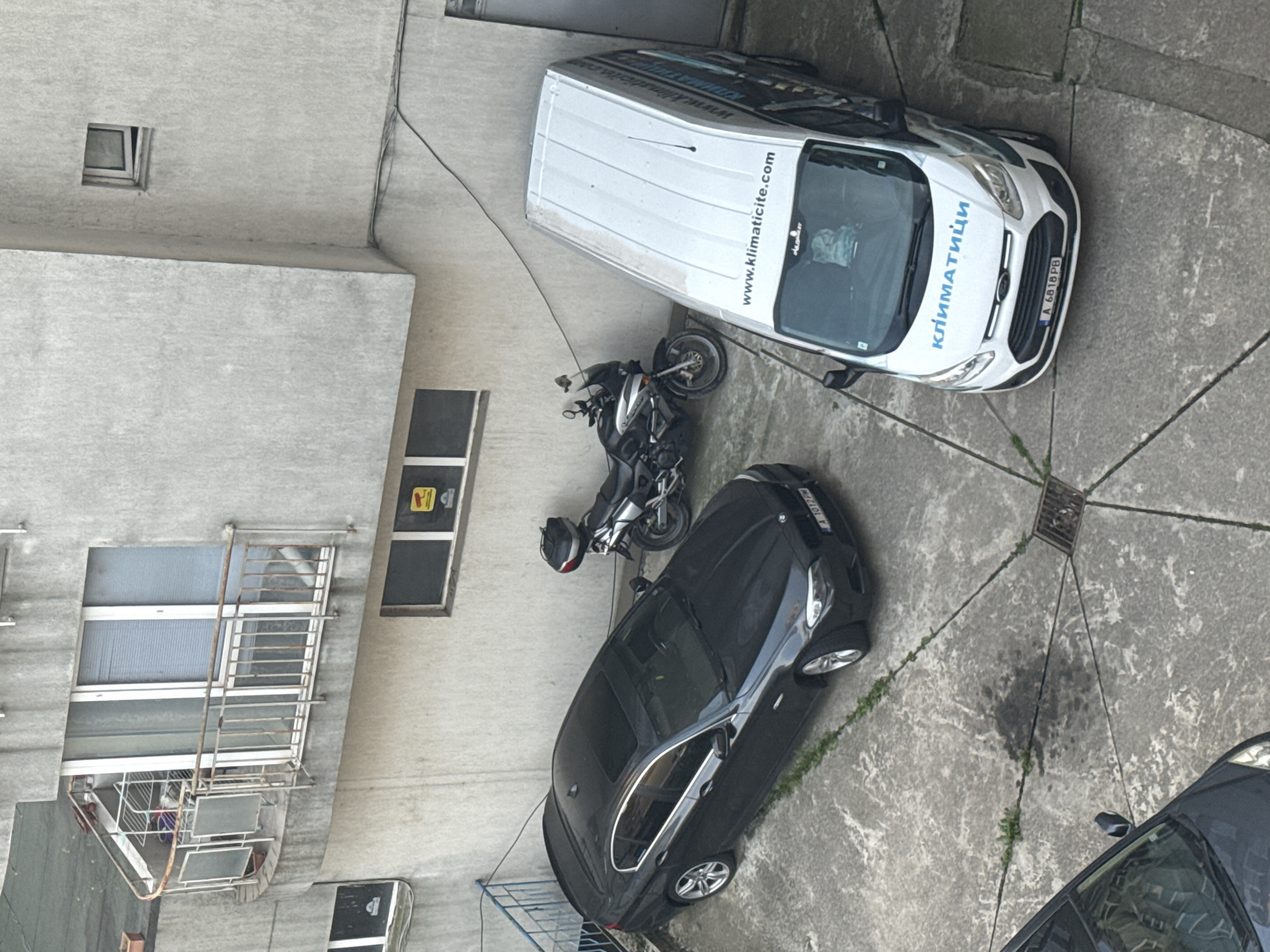 The image shows an overhead view of a courtyard in Burgas, Bulgaria. A white Ford van, bearing the markings "www.klimaticite.com" and "КЛИМАТИЦИ" along with the license plate "A 6618 PB," is parked near a motorcycle and a black car with the license plate "A 1077 PM." The area is paved with concrete, marked with cracks and some minor vegetation growing through them. Adjacent to the parking area is a building with windows, some having security bars. A drainage grate is visible in the pavement. There are no people visible in the image.