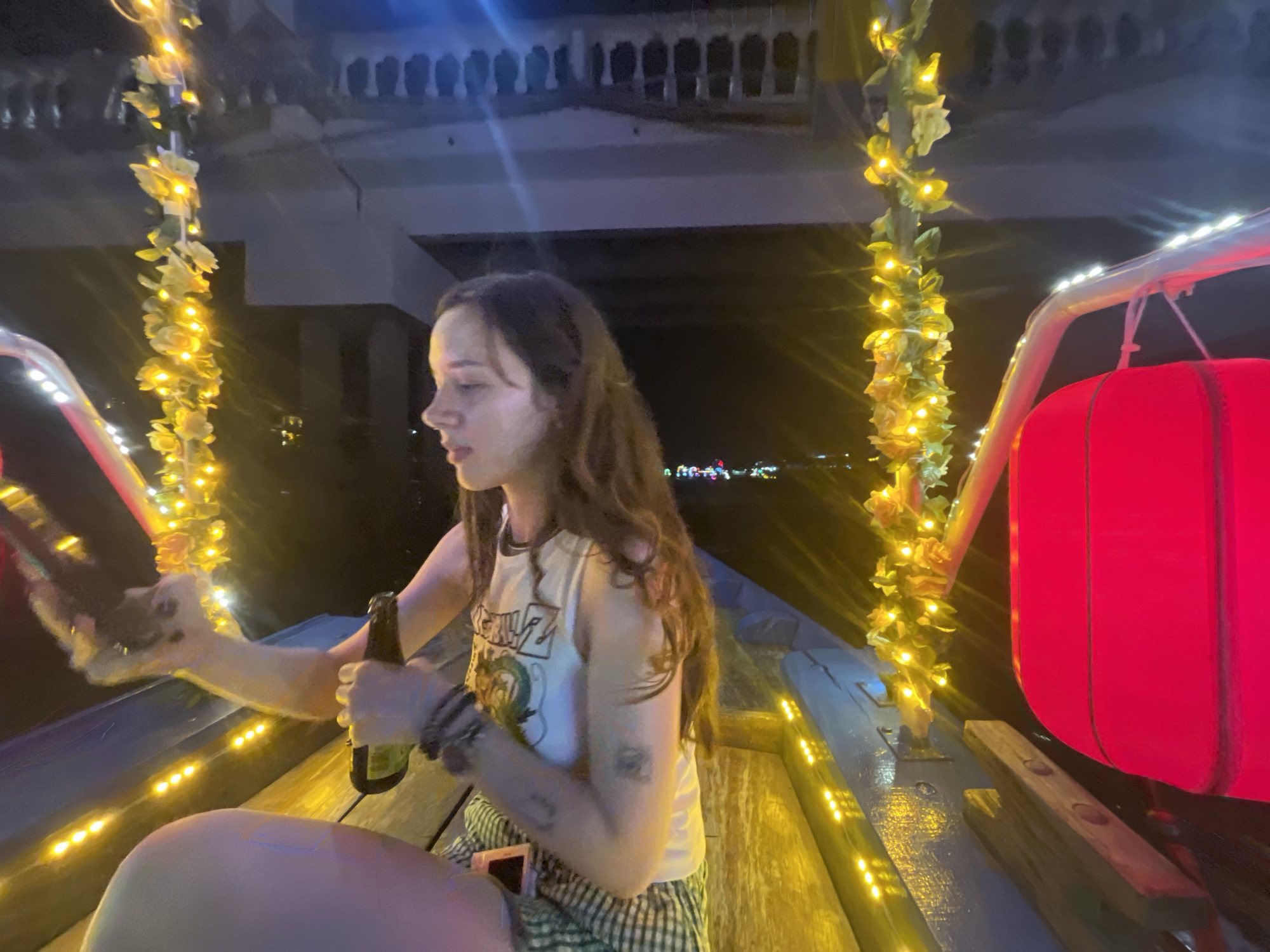 A young woman sits on the deck of a wooden boat illuminated by yellow string lights and faux ivy decorations. She is holding a bottle of beer in her right hand and appears to be looking to her left. The boat is adorned with a large red lantern on the right side. In the background, a bridge with ornate architecture is visible, and further in the distance, city lights twinkle. This scene is consistent with a boat tour on the Thu Bon River in Hoi An, Vietnam, at night.