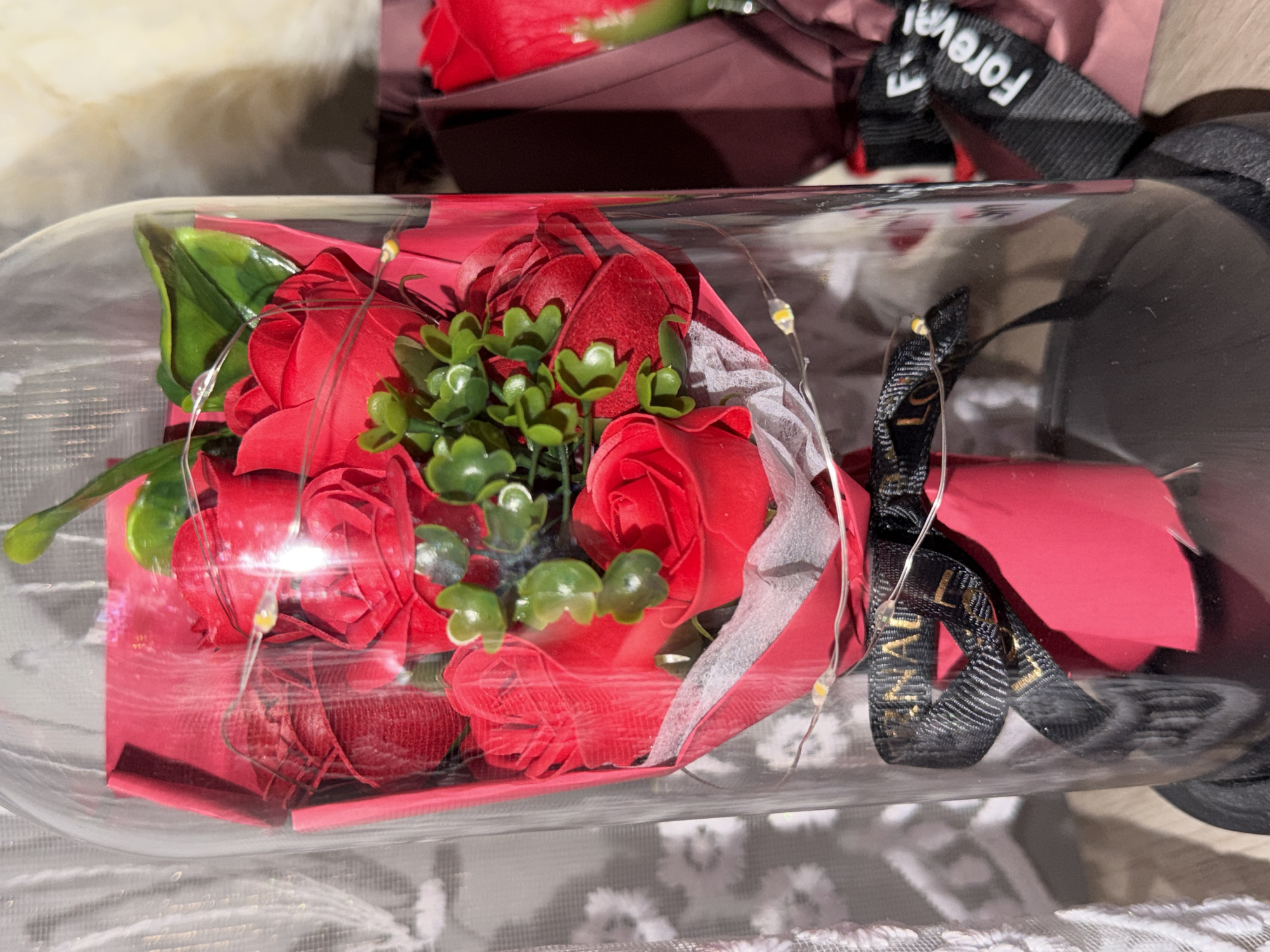 The scene depicts a clear glass dome containing a bouquet of artificial red roses, illuminated by a string of small LED fairy lights. The roses are wrapped in a combination of red paper and a crinkled white material. Small clusters of green artificial foliage are interspersed among the red blooms. A black ribbon with gold lettering, partially visible, is tied around the base of the bouquet within the dome. The glass dome rests on a dark, circular base. To the left, a white sheer curtain or lace fabric with a floral pattern is visible. To the right, a second, partially visible item consists of a single red artificial rose in dark reddish-brown packaging, secured with a black ribbon featuring white lettering that reads "Forever." The items are placed on a light-colored surface, possibly wood or laminate. The image provides no specific visual cues to determine a precise location within the city of Varna, Bulgaria, beyond being an indoor setting.