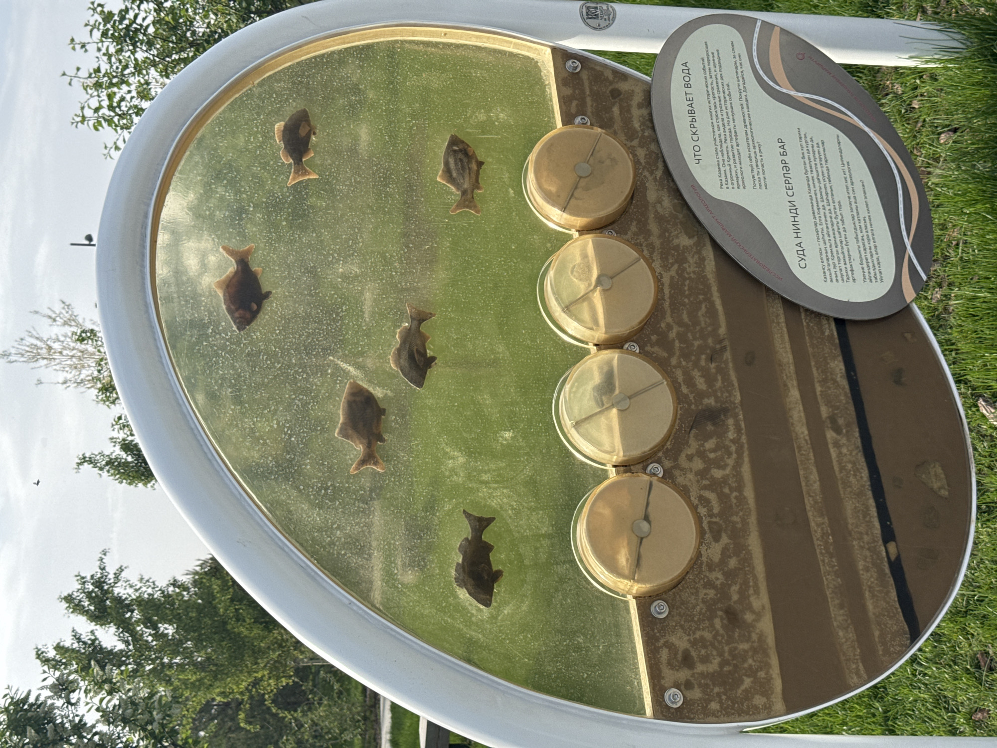 The image shows an outdoor informational exhibit, likely in a park setting. The exhibit consists of a circular display case with several fish-shaped objects embedded in a clear, possibly resin, surface. Four circular dials are adjacent to this display, presumably to be rotated to reveal hidden elements or information. A brown, oval sign is attached to the display, containing text in both Russian and what appears to be a Turkic language (possibly Tatar), suggesting the location is in Kazan, Russia, the capital of the Republic of Tatarstan. The sign describes the history of the Kazanka River and archaeological finds within it, inviting visitors to interact with the exhibit. The background reveals greenery and trees, suggesting a natural environment.