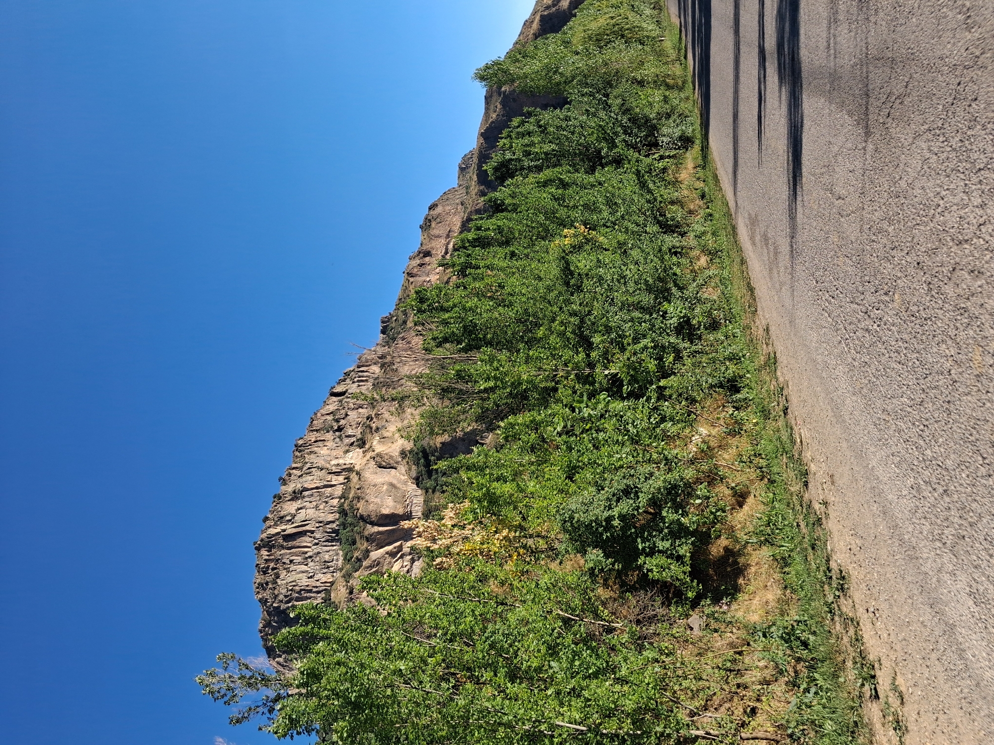 The image depicts a scenic view of a rugged, rocky cliff face covered in lush green vegetation. The cliff is steep and appears to be composed of layered sedimentary rock. The vegetation consists of dense bushes and trees, some of which have yellow foliage, suggesting either a particular type of plant or the season. The scene is bathed in bright sunlight, indicating a clear day, likely in the late morning or early afternoon due to the shadows cast by the cliff onto the adjacent road.
To the right of the cliff and vegetation, a paved road runs diagonally across the frame. The road's surface is asphalt, and its texture is visible, with small pebbles embedded in it. Long, dark shadows stretch across the road, originating from an unseen source, likely trees or poles off-frame to the left. The road appears to be empty of vehicles, suggesting a quiet, possibly rural, location.
The sky is a vibrant, cloudless blue, filling the upper left portion of the image. The overall impression is one of a natural landscape, perhaps a hiking trail or a scenic drive. The orientation of the image is such that the cliff face and road are seen from a tilted perspective, with the sky dominating the left side and the road the right.
The location context provided is Shatin, Armenia. This implies the image was taken in a mountainous or hilly region of Armenia. There is no visible text in the image. The scene evokes a sense of natural beauty and tranquility, with the grandeur of the cliff contrasted by the vibrant greenery and the man-made road.