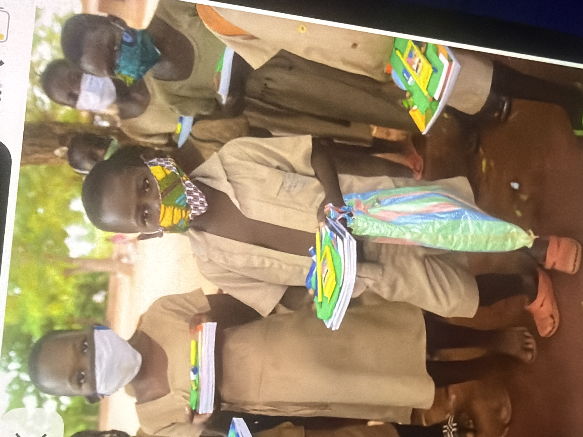 In an outdoor setting, possibly a schoolyard, a group of children are standing together. The child in the foreground, a boy, is wearing a tan school uniform and a colorful face mask. He is holding a bundle of notebooks and a plastic bag with multi-colored stripes. To his left, a girl also in a tan uniform and a white face mask holds a stack of notebooks. Behind them, other children are visible, some wearing face masks and school uniforms. The environment appears to be natural, with trees in the background. The ground is a reddish-brown color, suggesting soil or dirt. The overall scene depicts children receiving or holding school supplies.