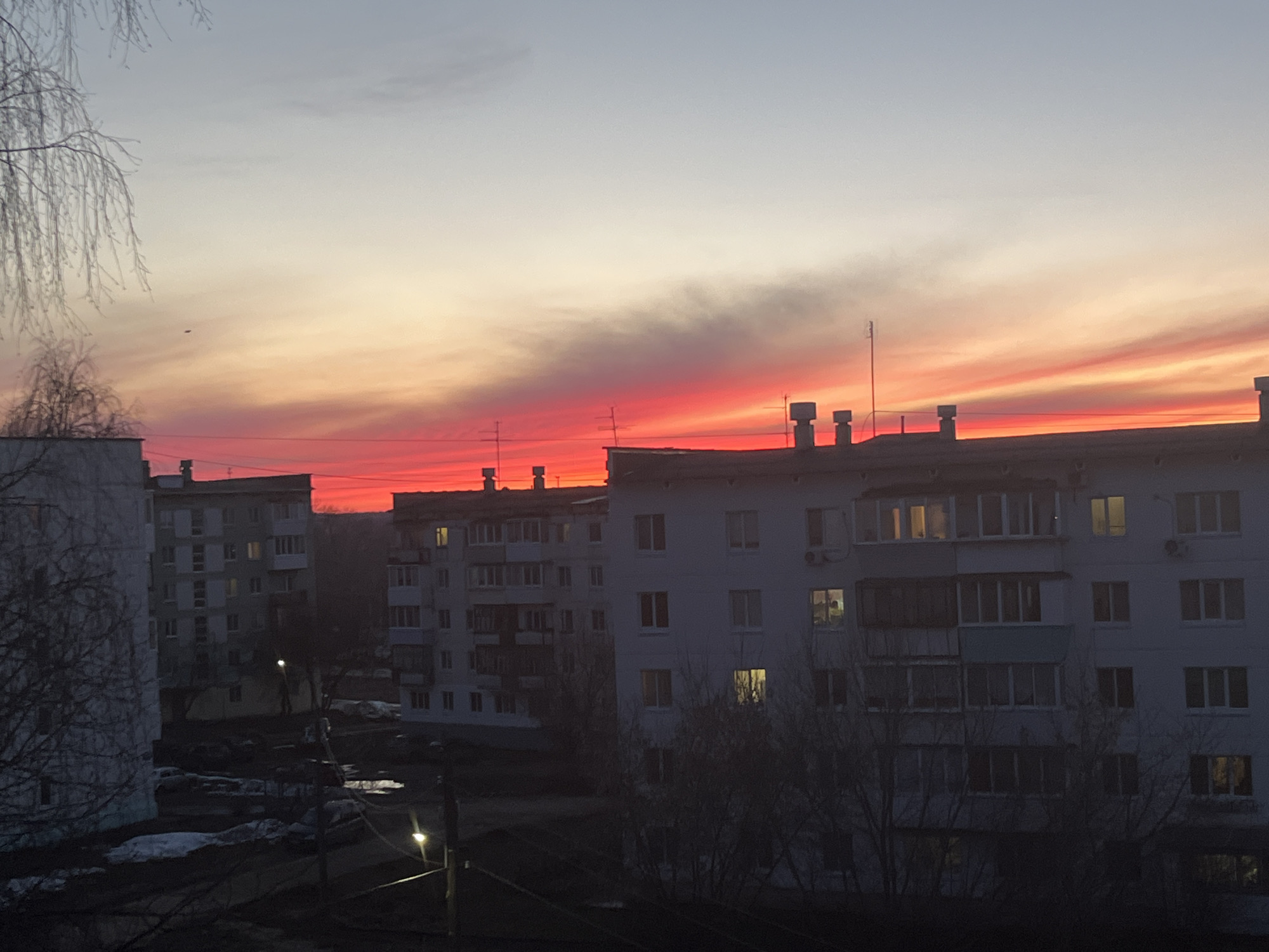 The image depicts a twilight or dawn scene over a residential area in Chaikovsky, Russia. Multiple Soviet-era apartment buildings dominate the view, silhouetted against a vibrant sky with hues of red, orange, and pale blue. Several windows are illuminated in the buildings, suggesting early morning or late evening. A few bare trees are visible in the foreground and scattered snow patches remain on the ground, indicating late winter or early spring. A street lamp illuminates a section of the foreground.