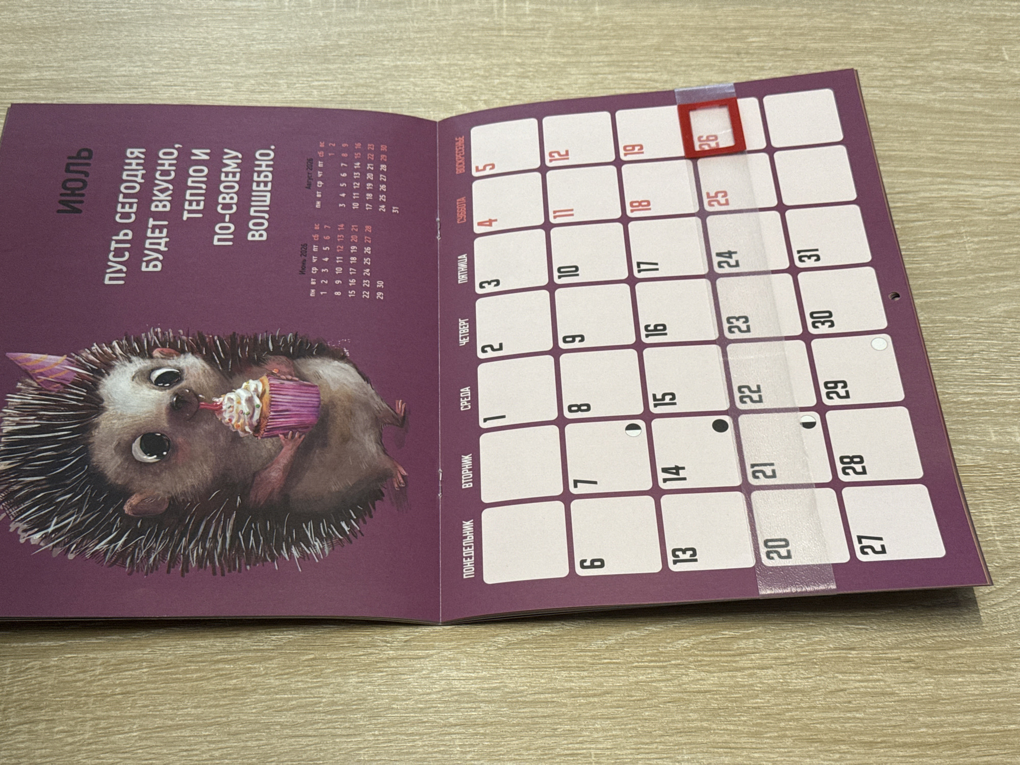 The image shows a calendar page for July, featuring an illustration of a hedgehog holding a cupcake. The calendar grid displays dates from July 1st to July 31st. The page is purple and has Russian text, including the month "ИЮЛЬ" (July) and a motivational quote. Below the illustration and text is a grid layout of the days of the week: ПОНЕДЕЛЬНИК (Monday), ВТОРНИК (Tuesday), СРЕДА (Wednesday), ЧЕТВЕРГ (Thursday), ПЯТНИЦА (Friday), СУББОТА (Saturday), and ВОСКРЕСЕНЬЕ (Sunday). The dates are numbered within their respective day boxes. A red border highlights the 26th of July. The calendar is attached to a wall with a wood-grain texture. The location is identified as Tomilino, Russia.