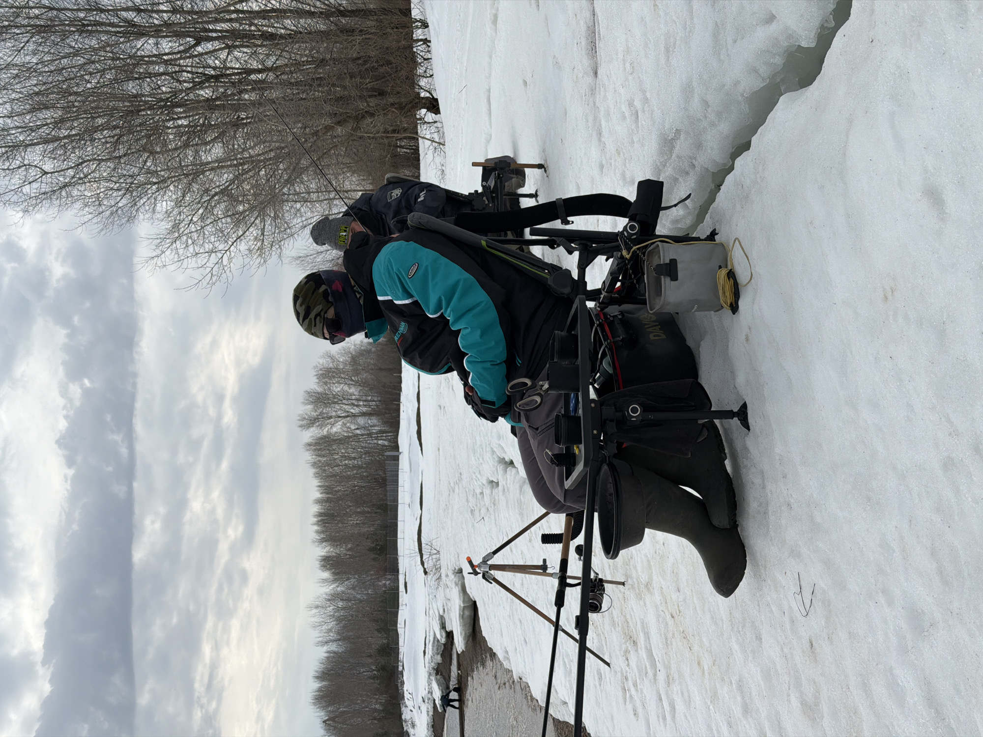 Two individuals are ice fishing in a snowy environment, likely in Noviy, Russia, given the prompt. They are dressed in winter gear, including hats, jackets, and boots. One individual has a black jacket with teal accents and a camouflage hat. They are seated with various fishing equipment around them, including a fishing rod on a tripod, a tackle box, and possibly a sled or chair designed for ice fishing. Patches of snow cover the ground. The trees in the background are bare, indicating it is likely late winter or early spring. The sky is overcast.