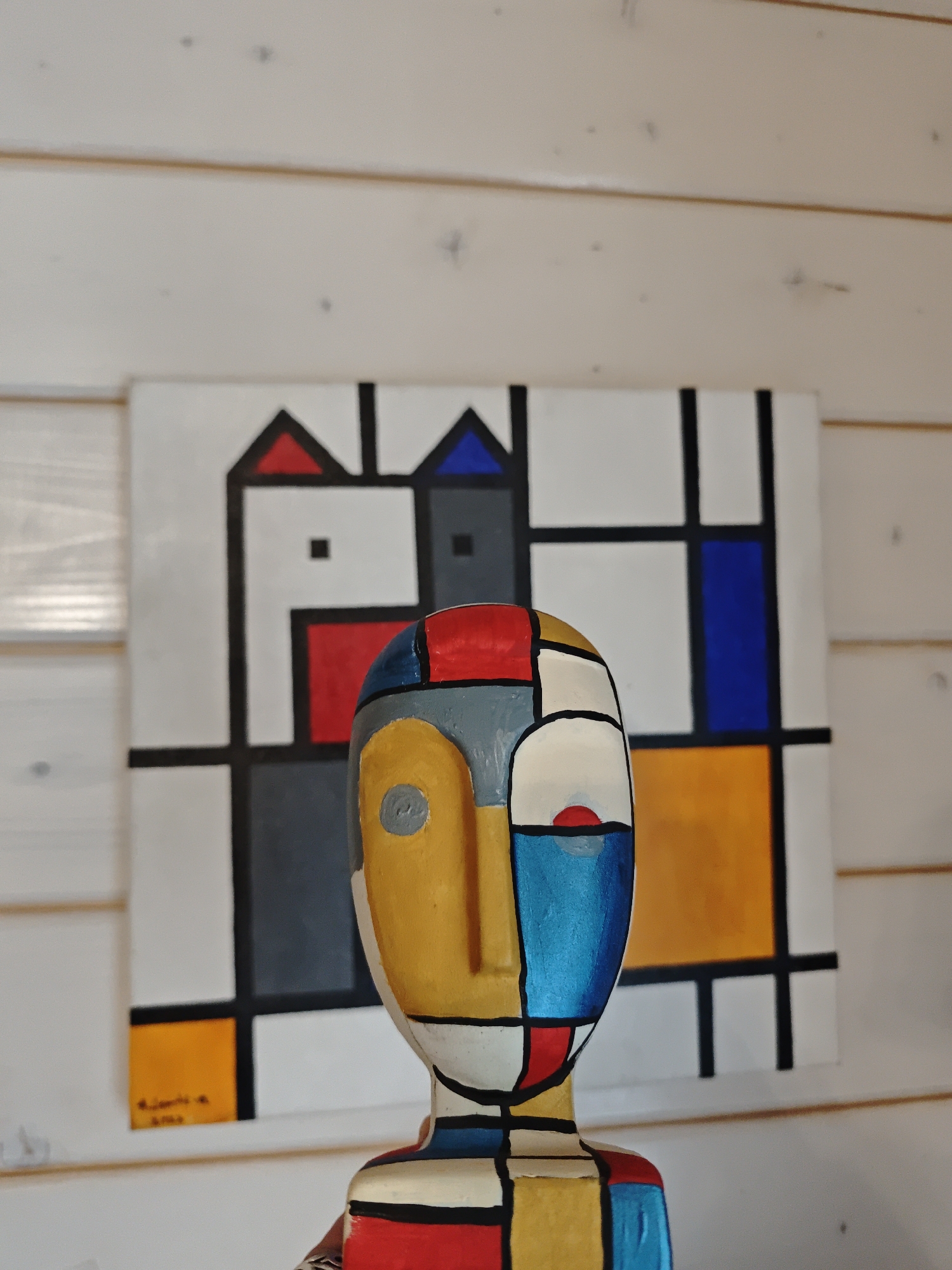 The image shows a wooden sculpture of a face with a Mondrian-style paint job. It is segmented into various geometric shapes and painted in red, blue, yellow, white, gray and black. The sculpture is standing in front of a painting. This painting is also in the style of Mondrian, featuring a grid of black lines and filled with geometric shapes in similar colors. The background is a wall made of horizontal wooden planks painted white. The location is in Свистуха (кузяевский С/о), Russia.