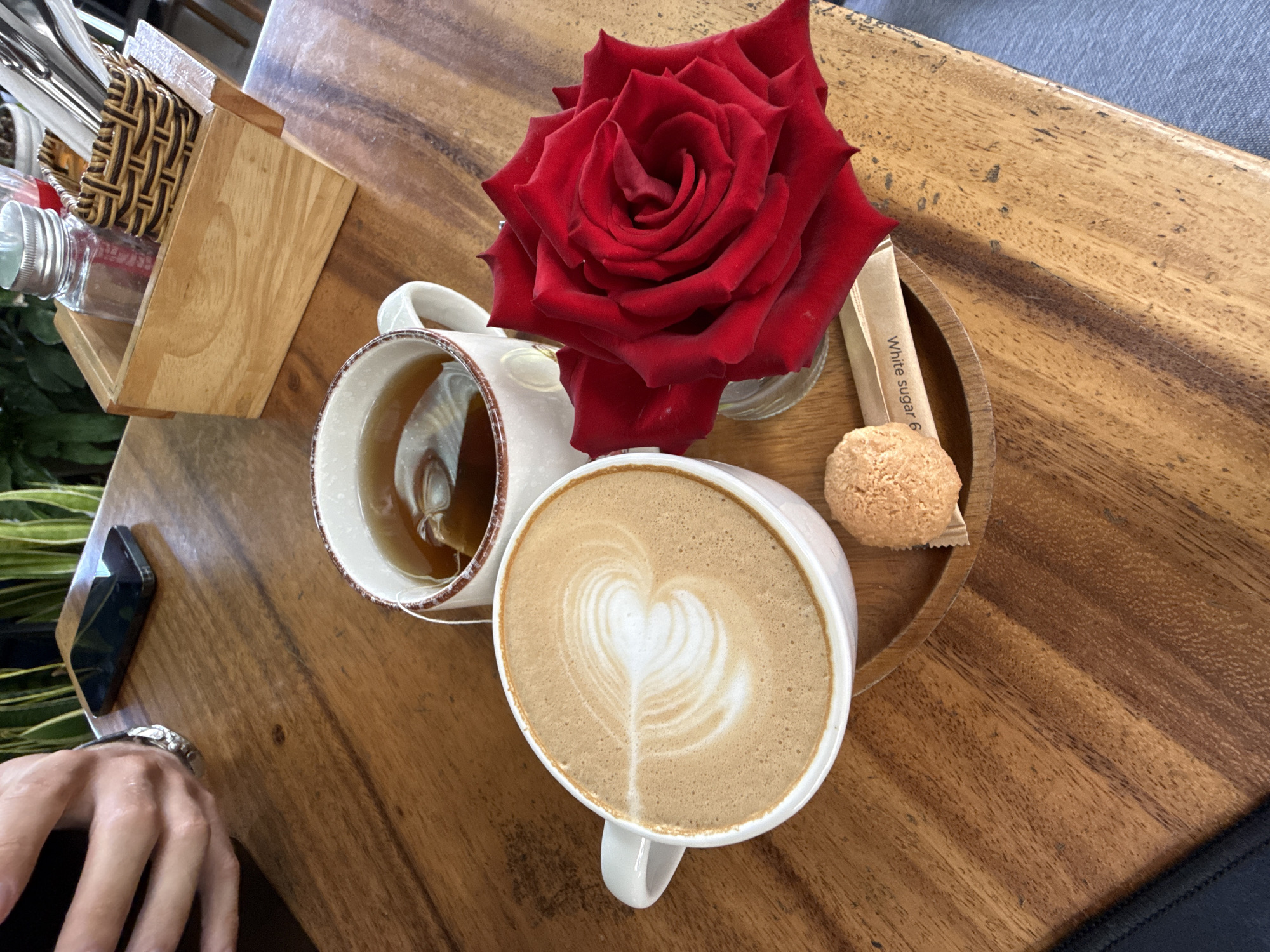 A top-down shot shows a wooden table surface with a cup of tea, a latte with a heart design, a rose in a small vase, a sugar packet, and a small cookie on a circular wooden tray. To the left, there's a wooden container with utensils and a bottle. A phone sits next to the tea, near a hand with a watch. The scene is likely set in a cafe, possibly in Da Lat, Vietnam, given the local aesthetics and coffee culture often found there.