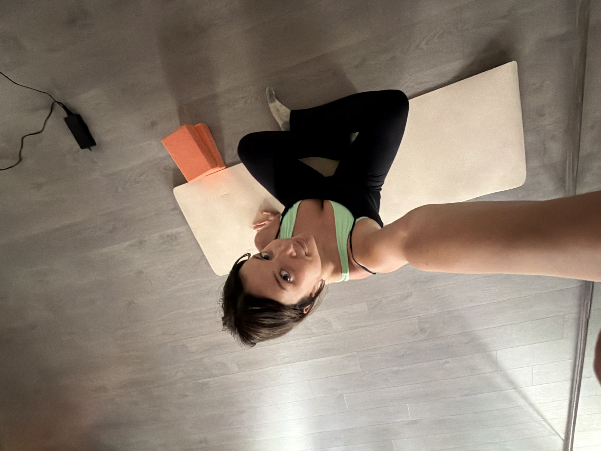 A woman in a light green sports bra and black leggings is sitting on a yoga mat on a light gray laminate floor. She is holding a phone in her right hand, taking a selfie. Two orange yoga blocks are stacked on top of the yoga mat to her right. A black power adapter with a cord is on the floor to the left of the yoga blocks. The scene appears to be indoors, likely in a residential setting within Vracar Municipality, Serbia.