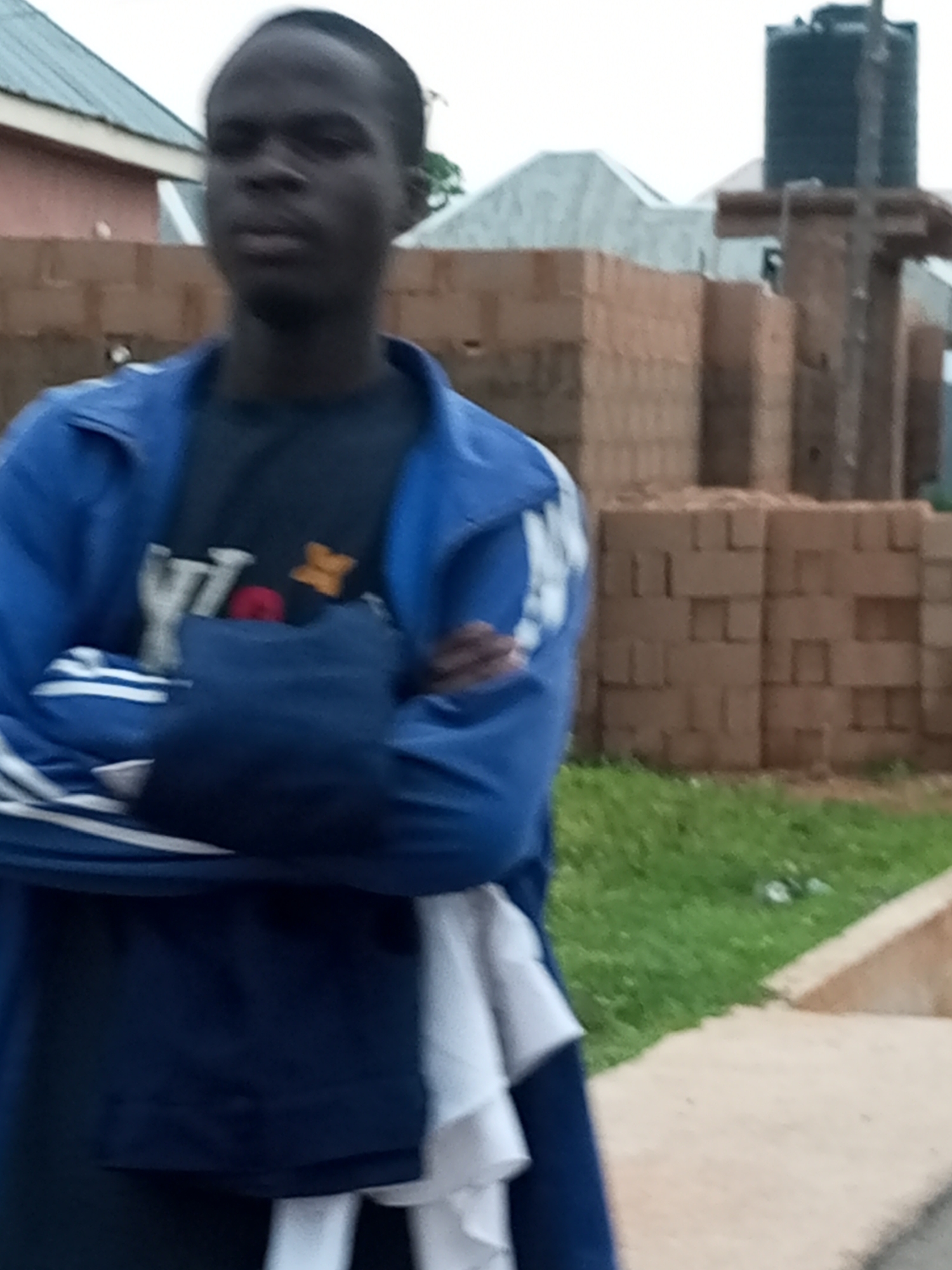 In an outdoor scene in Unknown, Unknown, a young Black male stands with his arms crossed. He is wearing a blue tracksuit with white stripes on the sleeves and a dark t-shirt underneath, which has a graphic design featuring the letters "Y" and "X" along with a red circular shape and an orange star. The man's expression is neutral as he looks towards the camera. Behind him, stacks of reddish-brown bricks are visible, suggesting a construction site or material storage. A dark water tank is perched on a structure in the background, along with parts of buildings with white roofs. Green grass covers the ground to the right of the man, bordering a beige walkway or path. The overall lighting indicates an overcast or daytime setting.