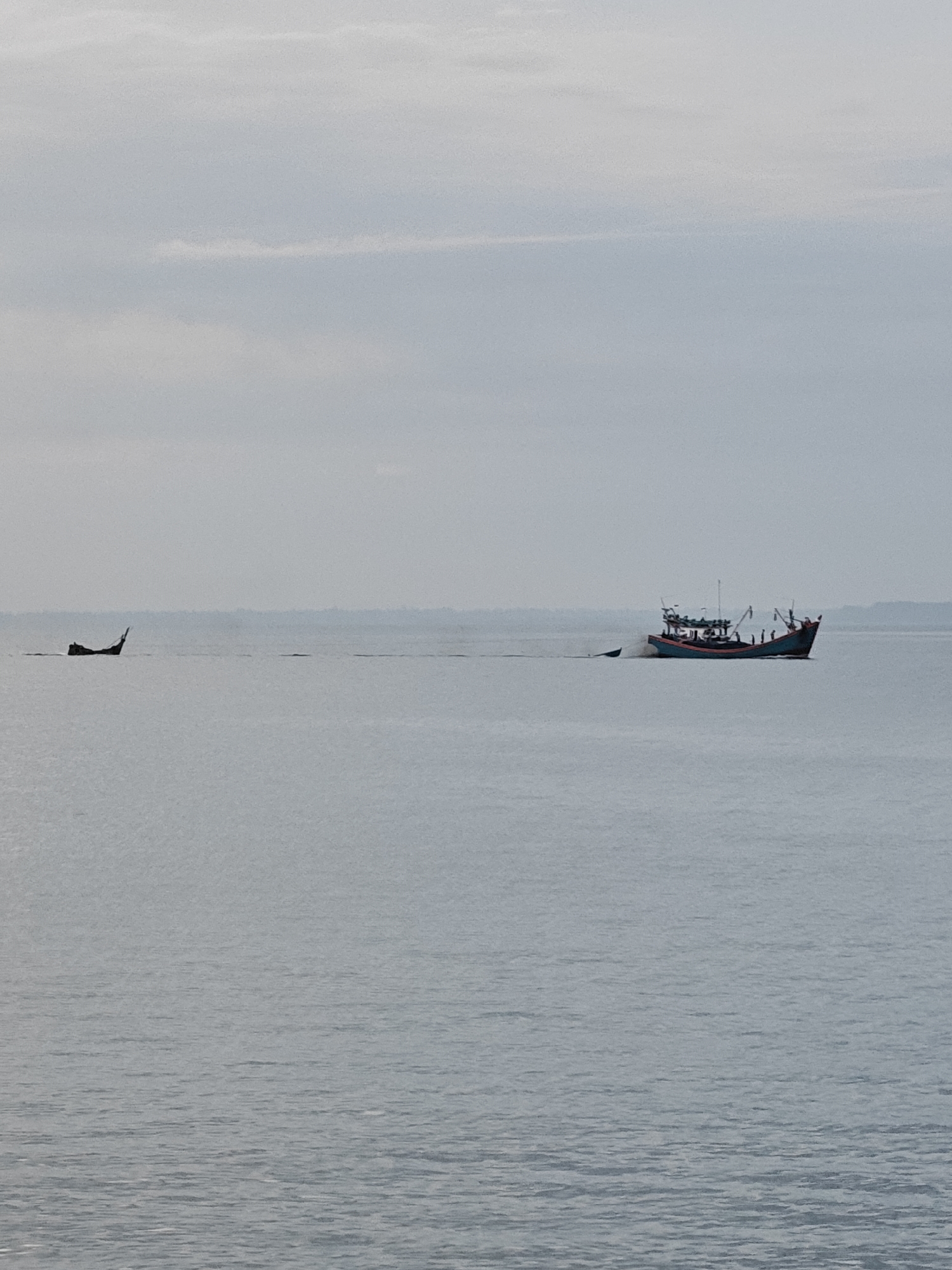 The image depicts a vast expanse of calm, grayish water under an overcast sky. In the middle distance, two boats are visible. The boat on the right is a larger, colorful fishing vessel with multiple decks and rigging, moving from left to right. The boat on the left appears to be a smaller, darker vessel, possibly a longtail boat, also moving towards the right, leaving a visible wake. The horizon is hazy, suggesting a distant coastline or low-lying land. The overall scene conveys a sense of quiet activity on the water. This is the sea off the coast of Lhokseumawe, Indonesia.