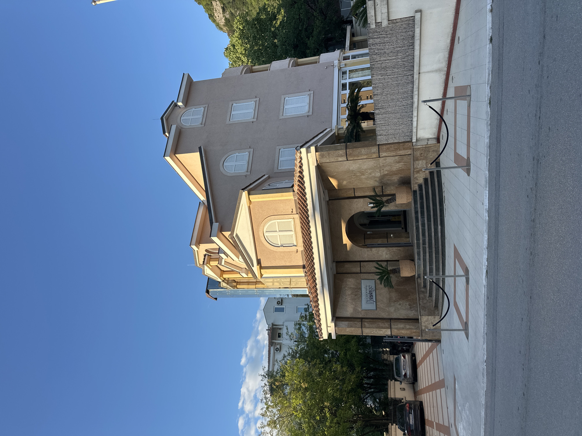 The image shows the front of a multi-story building in Budva, Montenegro, likely a hotel named "Hotel Voyage." The building has a light brown facade with white window shutters and is partially obscured by a tree in the foreground. Steps lead up to the building's entrance. The street is visible in the lower right of the image, along with parked cars. The sky is clear and blue.