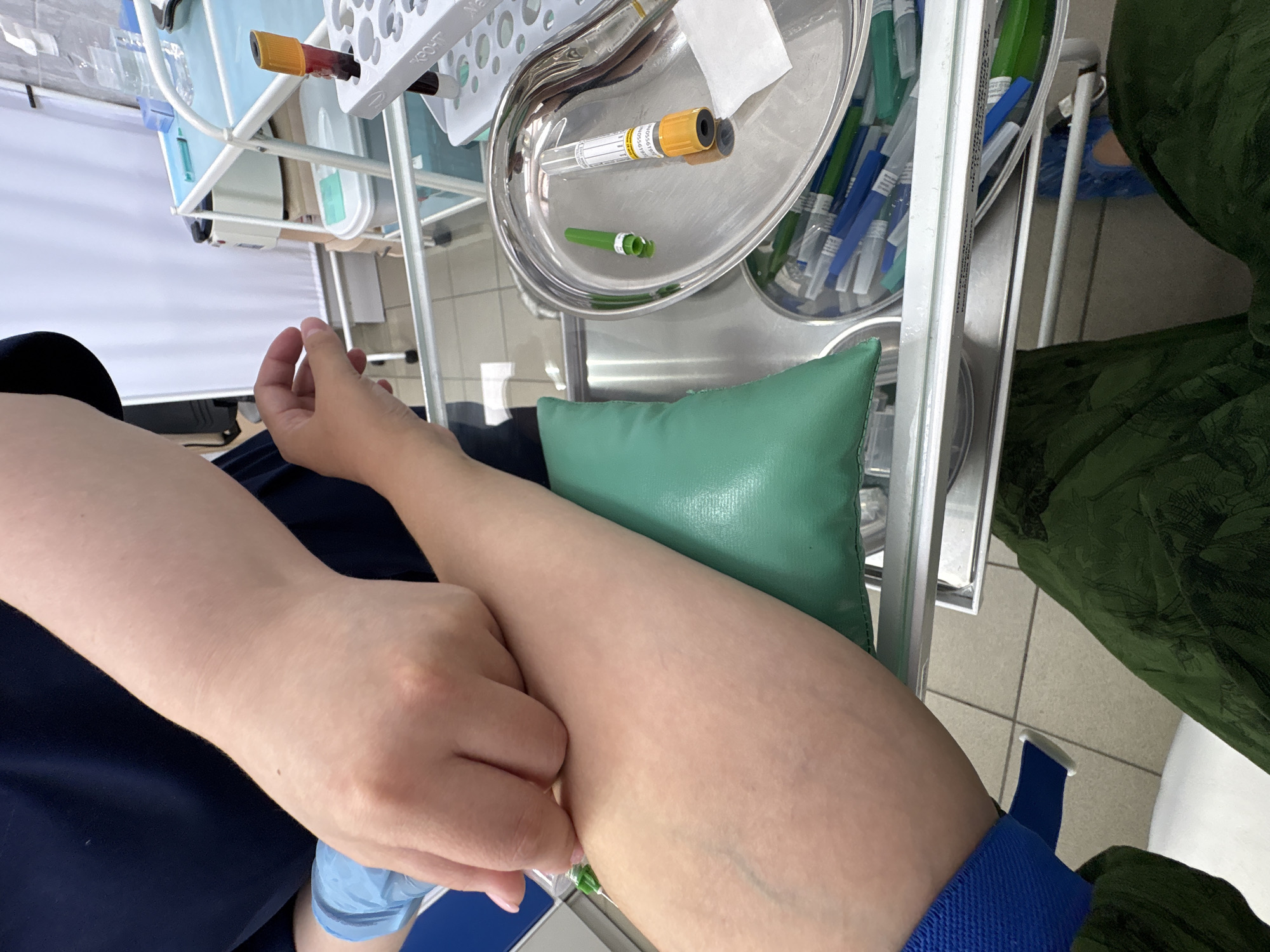 The image depicts a close-up view of a blood draw procedure. A person's forearm, supported by a green cushion, is visible. A healthcare professional, wearing blue gloves, is inserting a needle into the vein. A tourniquet is applied to the upper arm. In the background, medical equipment and supplies are present, including a tray with blood collection tubes and needles. The scene appears to be in a medical setting. There is no indication of the specific location within Birobidzhan, Russia.