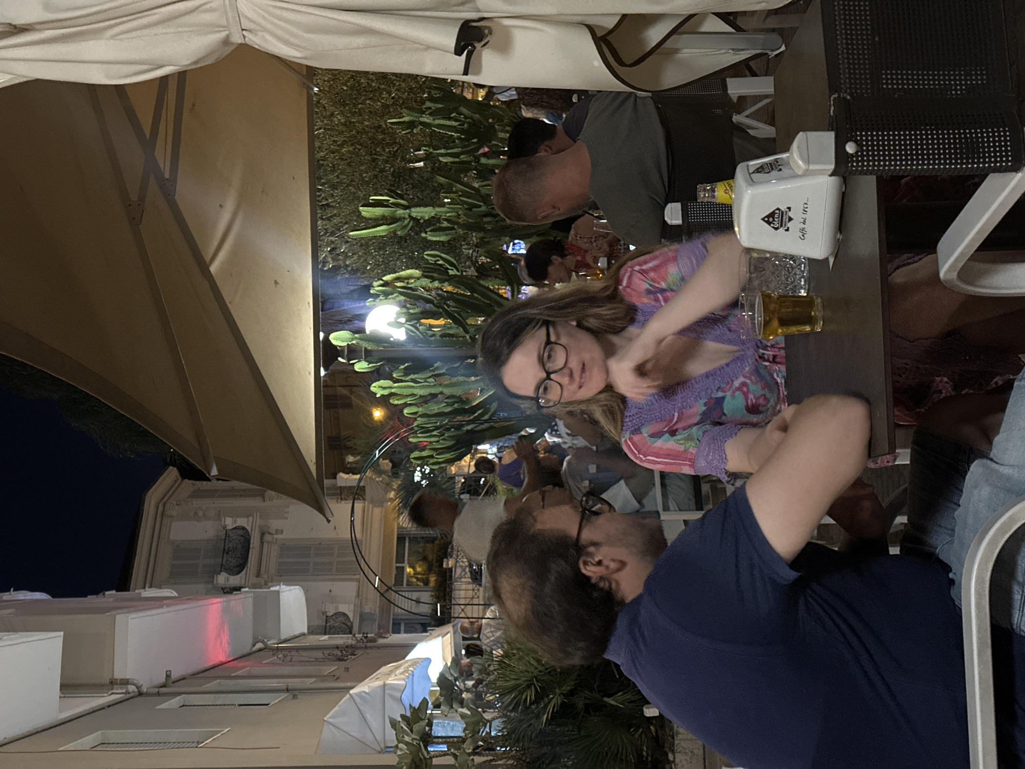 The image depicts an outdoor seating area of a cafe in Ospedaletti, Italy, at night. Two people, a man and a woman, are seated at a table in the foreground, appearing to be in conversation. The man, on the left, has dark hair and wears a dark blue polo shirt. He is looking to his left. The woman, on the right, has lighter hair and wears glasses and a purple and pink patterned top. She is looking down and to her left.
On the table are a glass of beer, an empty glass, a napkin dispenser with "Giena Caffè dal 1869..." printed on it, and a bottle of Schweppes. Behind the couple, other patrons are visible in the background, along with large potted cacti and foliage. Buildings with architectural details, typical of Italian coastal towns, are seen in the distance, illuminated by artificial light. A large beige umbrella provides shade over the seating area. The overall atmosphere suggests a relaxed evening at a cafe.