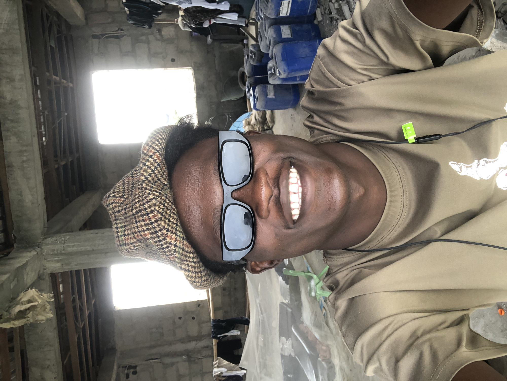 The image shows a close-up selfie of a Black male wearing a brown, plaid flat cap and dark sunglasses. He is smiling and looking directly at the camera. He is wearing a tan t-shirt with black earbuds around his neck. The background appears to be an unfinished or abandoned building with exposed concrete columns and ceiling beams. There are two large windows with bright light coming through them. To the right of the man, there are stacks of blue plastic drums and some clothing hanging on a rack. To the left, there is a light-colored tarp or sheet draped over something, and a green chair. The overall environment suggests a construction site or a storage area in a less developed part of Ado, Nigeria.