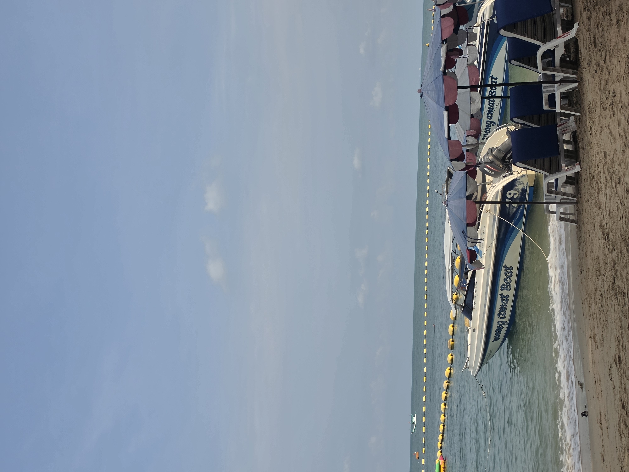 The image captures a coastal scene in Pattaya City, Thailand, featuring two speedboats beached near the shoreline. The boats are painted in white with blue stripes and bear the name "wong amat Boat" in blue lettering along their sides. Above each boat, there's a set of beach umbrellas with alternating white, red, and blue panels. The foreground shows beach chairs, arranged adjacent to the boats. A string of yellow buoys extends into the water, marking off a swimming area. The beach itself is sandy. The sea water is a clear greenish-blue. In the background, the sky is a light blue color.