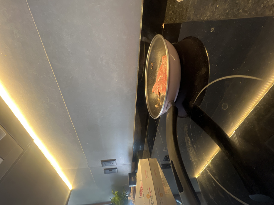 The scene depicts a contemporary kitchen environment located in León, Mexico. In the mid-ground, a light pink frying pan containing several slices of raw or lightly seared red meat, seasoned with visible spices, rests on a black induction cooktop. To the left of the pan, a white Krispy Kreme box, partially showing the word "RECAUDACIÓN," is present on the dark, reflective countertop. The background consists of a dark gray tiled or paneled backsplash wall with two dark electrical outlets mounted. Above the wall, a linear warm-toned LED light strip illuminates the area. A hint of green foliage from a plant is visible on the far left. No individuals are present in the image.