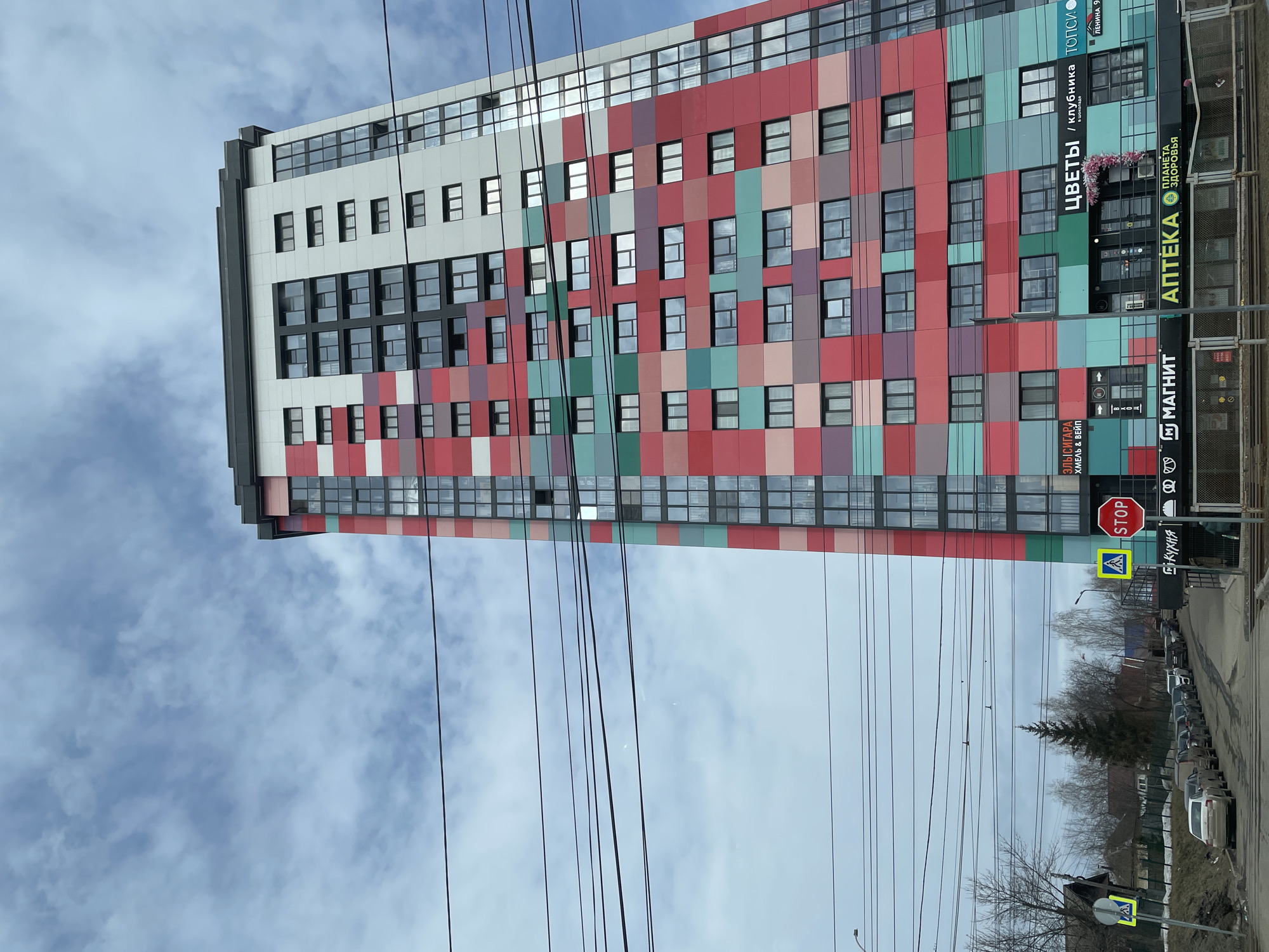 Here is a description of the image:
The image shows a tall, colorful building in Izhevsk, Russia. The building has a grid-like pattern of red, pink, green, and gray panels. There are windows on each panel, and some of the windows are reflecting the sky. The top of the building is white with black windows. On the ground level of the building are several businesses, including a flower shop ("ЦВЕТЫ"), a pharmacy ("АПТЕКА"), and a Magnit grocery store. There is a stop sign and a pedestrian crossing sign in front of the building. Power lines run across the image, obscuring some of the view. The sky is cloudy. Cars are parked along the street.