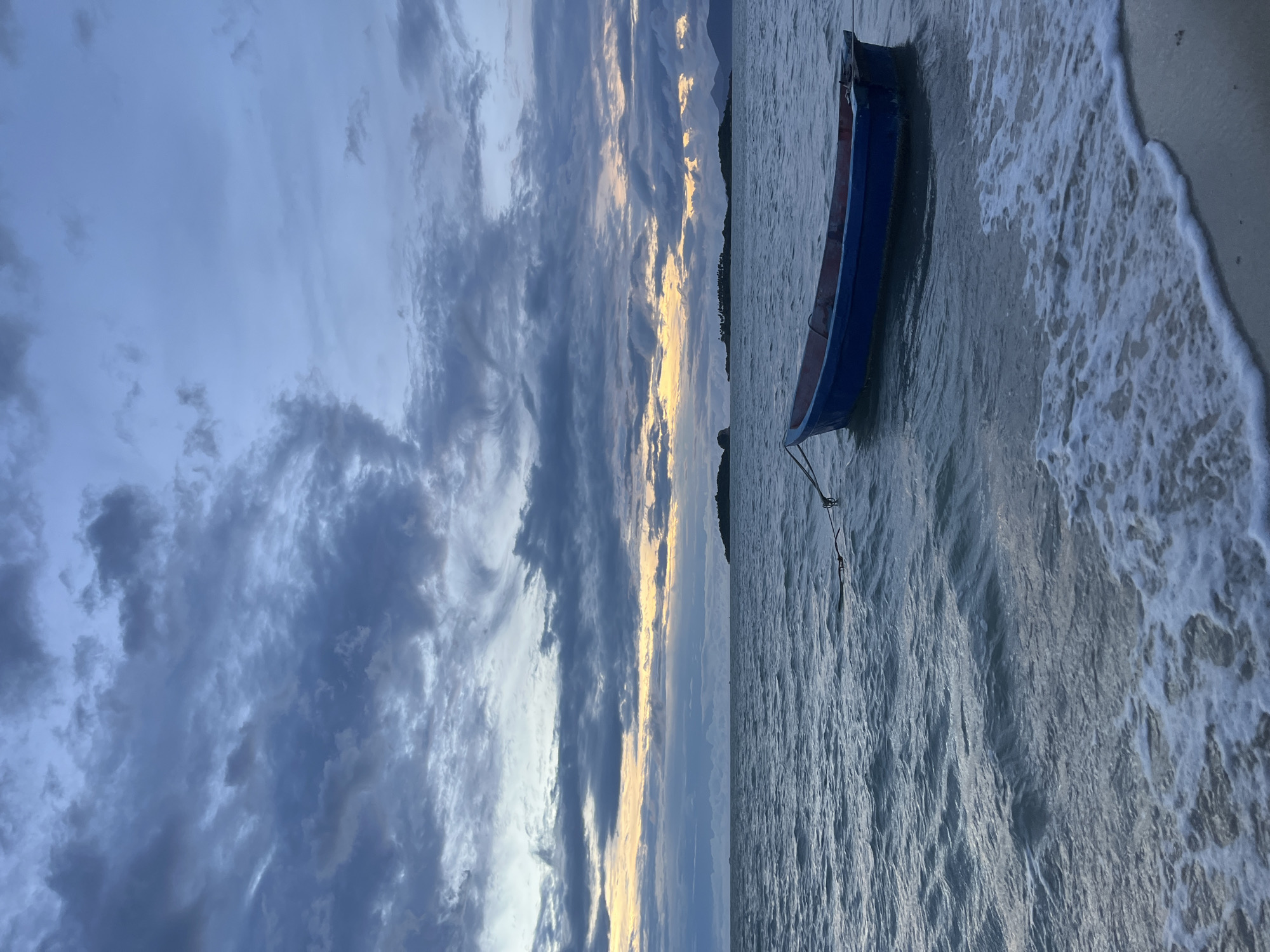 A blue boat with a red interior is tethered to an unseen anchor in the gentle waves of the sea. The boat is situated close to a sandy shore, where small waves with white foam are washing onto the beach. In the distance, several islands are visible, some with sparse vegetation and others appearing as dark silhouettes against the horizon. The sky above is filled with dramatic clouds in shades of grey and blue, with streaks of soft yellow and orange light peeking through, suggesting either sunrise or sunset. The overall scene evokes a sense of tranquility and natural beauty, characteristic of a coastal setting. The location is identified as Baan Plai Laem, Thailand.