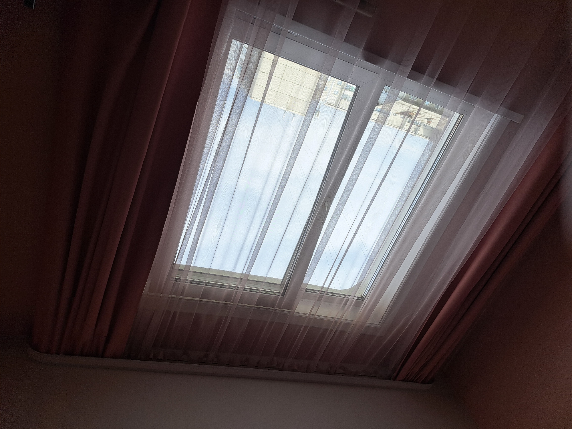 The image shows a window with sheer white curtains layered in front of thicker, rose-colored drapes. Through the window, a partially visible building with a tiled roof and multiple windows can be seen. The sky above appears to be partly cloudy. The scene suggests an indoor environment looking out onto a residential or urban area. No people or actions are depicted. The lighting indicates it is daytime.