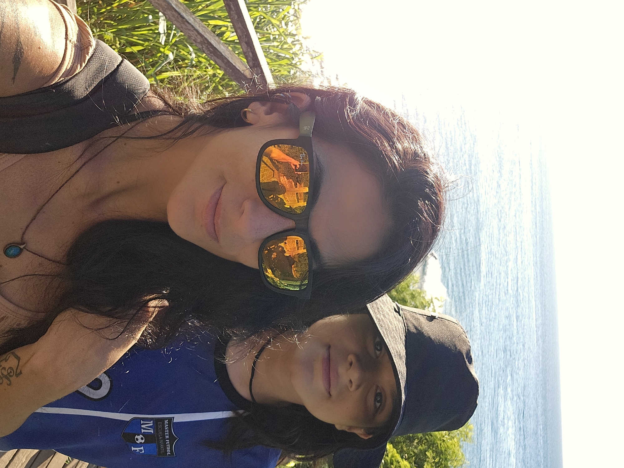 A sunlit selfie shows two individuals positioned close together, likely a mother and child, with an ocean backdrop, likely in Armação Dos Búzios, Brazil. The adult, located higher in the frame, has long dark hair, sunglasses that reflect a wooden structure, and a light complexion. She wears a necklace with a turquoise stone and has a tattoo on her arm. The child wears a dark baseball cap and a blue soccer jersey with a "MASTER FÚTBOL ESCUELA INFANTIL" logo. Both face the camera, sharing a similar complexion. The background features a pale blue ocean and bright sunlight, with hints of green vegetation. A weathered wooden rail is visible above the adult's head.