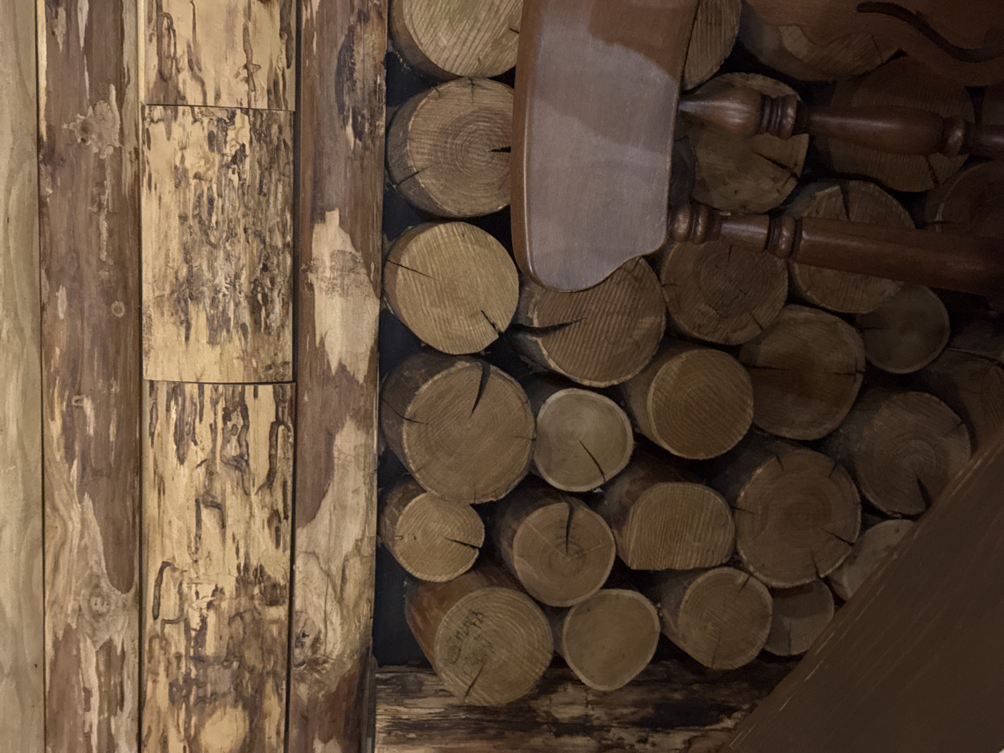 The image shows a close-up view of a rustic fireplace, constructed with logs and wooden planks. The logs are stacked neatly, forming a wall behind a portion of a wooden chair, which is partially visible in the foreground. The texture of the wood is rough and varied, with visible grain patterns and natural imperfections. The lighting is dim, casting shadows and highlighting the textures of the wood. The scene evokes a cozy, natural aesthetic, likely found in a cabin or a rustic interior setting. There are no people or actions depicted in the image. The location is not specified, and no information is provided about Palermo, Italy.